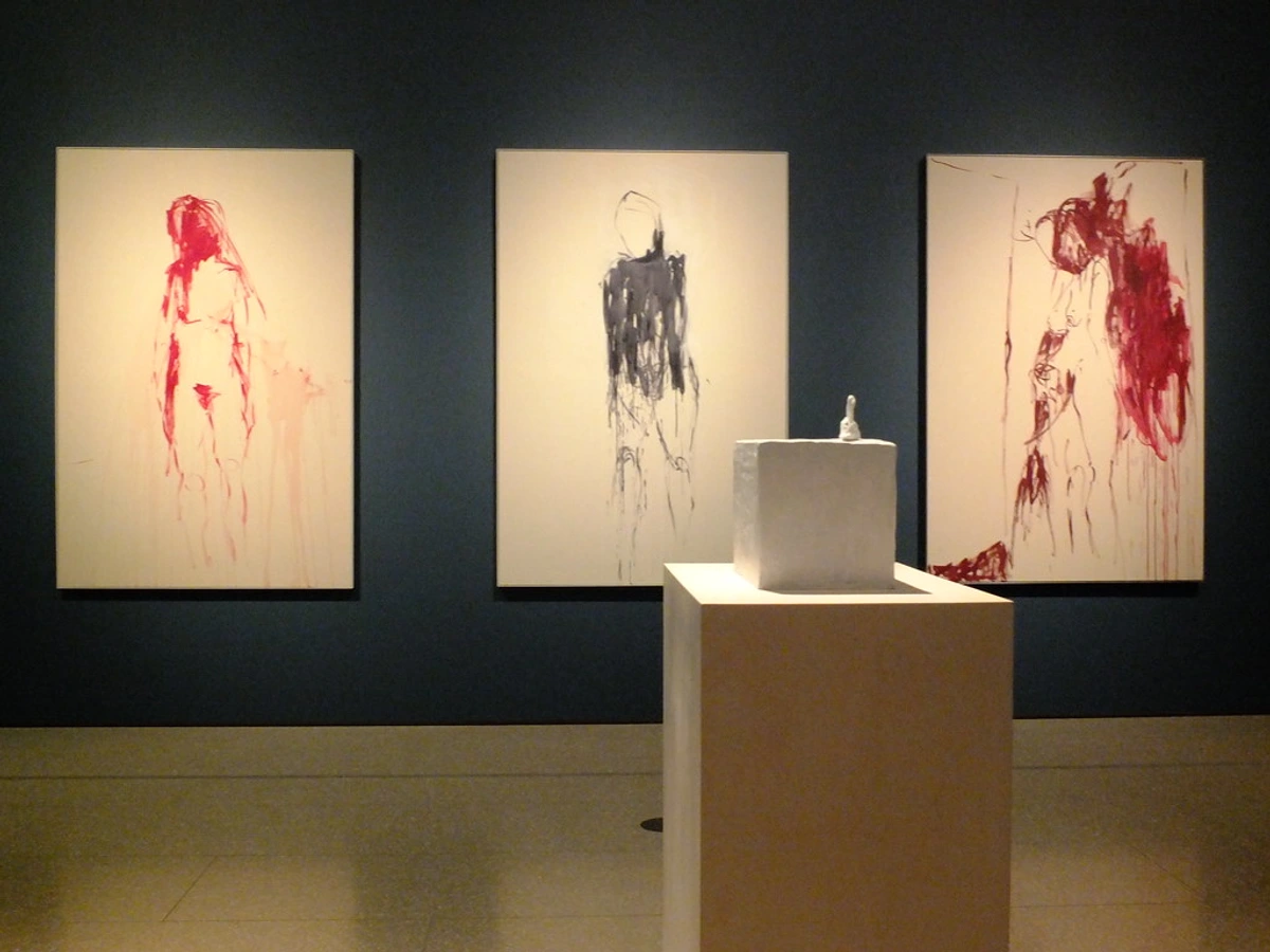 Tracey Emin triptych artwork featuring three painted figures in red and black on white canvases, displayed in a gallery with a small sculpture on a pedestal.