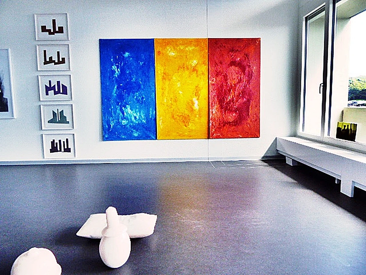 Triptych painting with blue, yellow, and red panels in a modern art gallery, alongside framed geometric art and abstract sculptures.