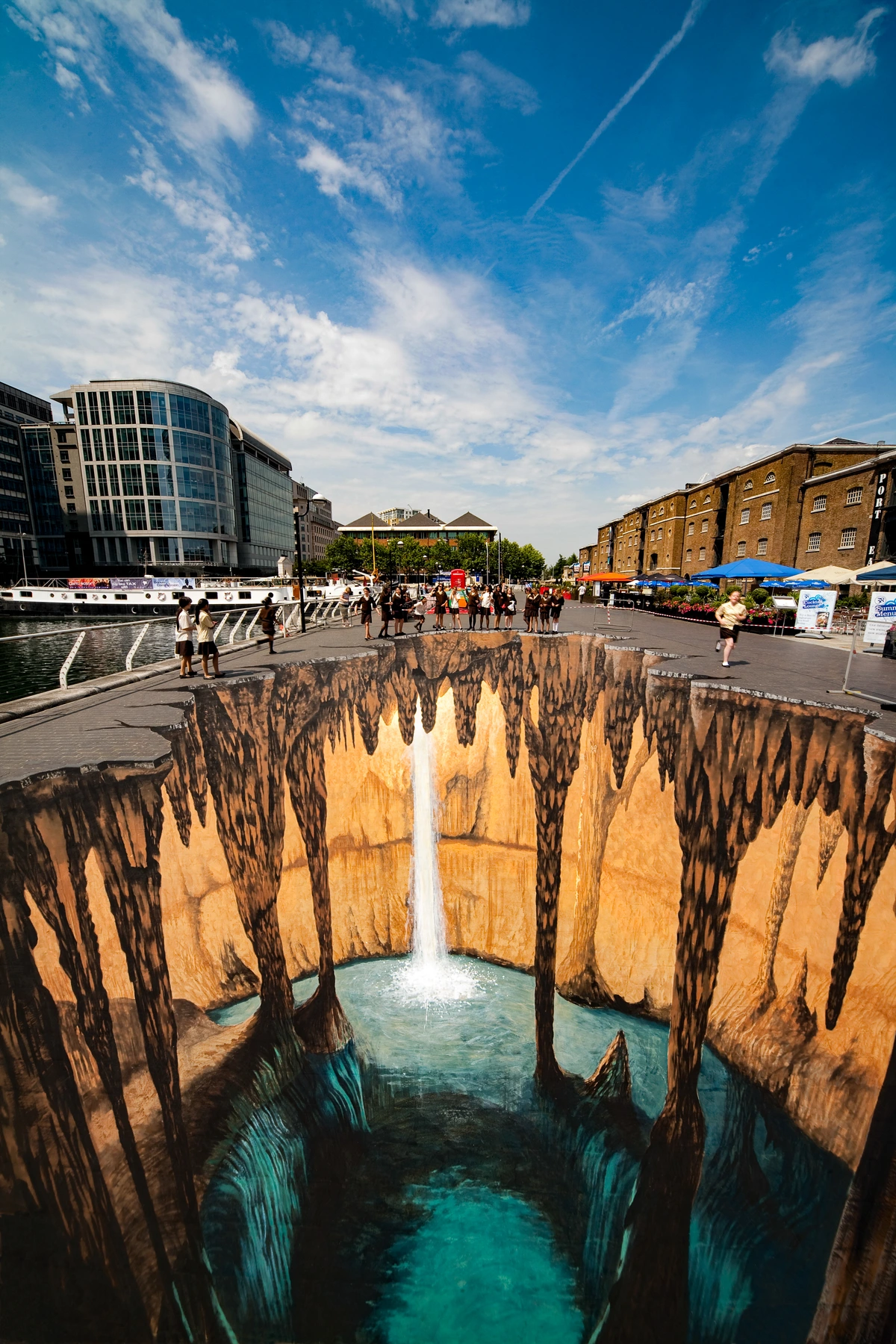 3D anamorphic street art depicting a cave with a waterfall, viewed by a crowd of people in London.