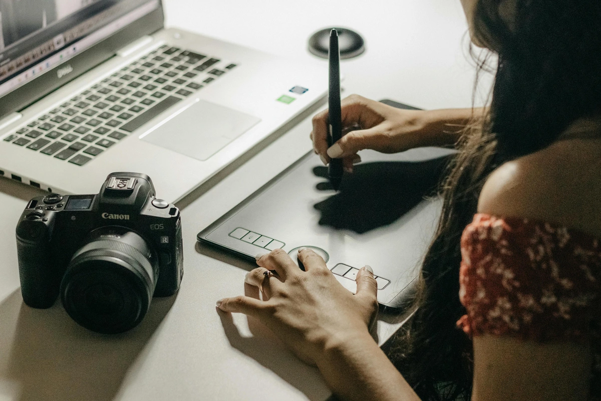 Woman using a digital tablet for creating art and taking notes in a creative workspace with a professional camera and laptop for documentation. Ideal for discussions on digital art production and critical reception studies.