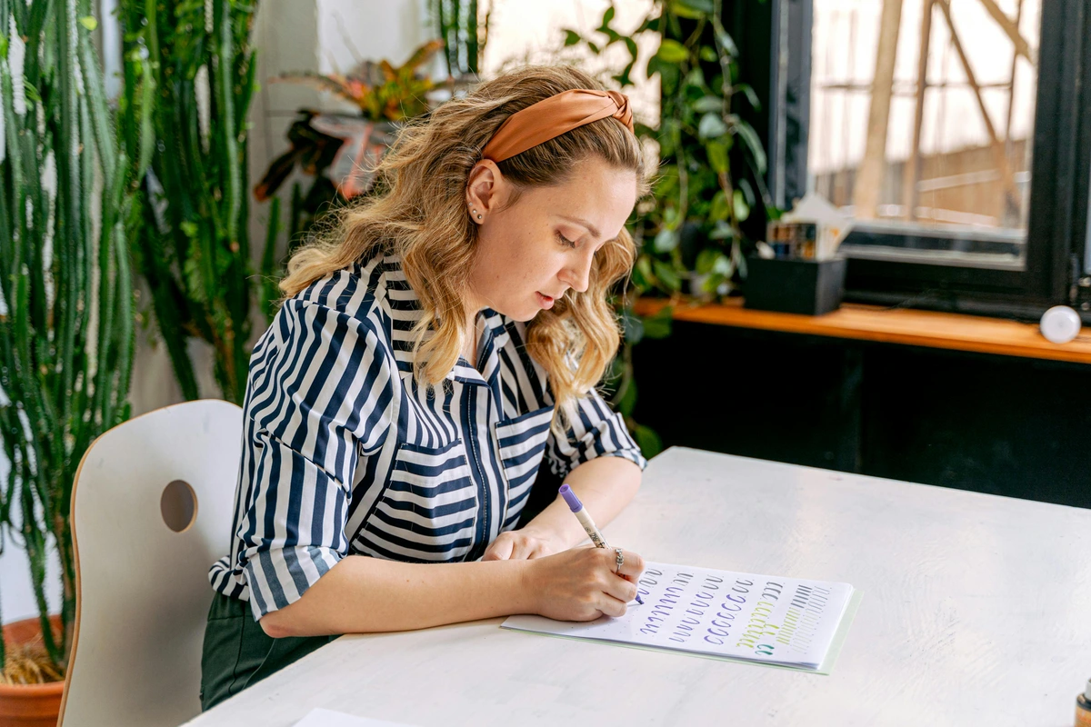 Woman practicing hand lettering at an art school desk, focused composition with artistic tools in natural light