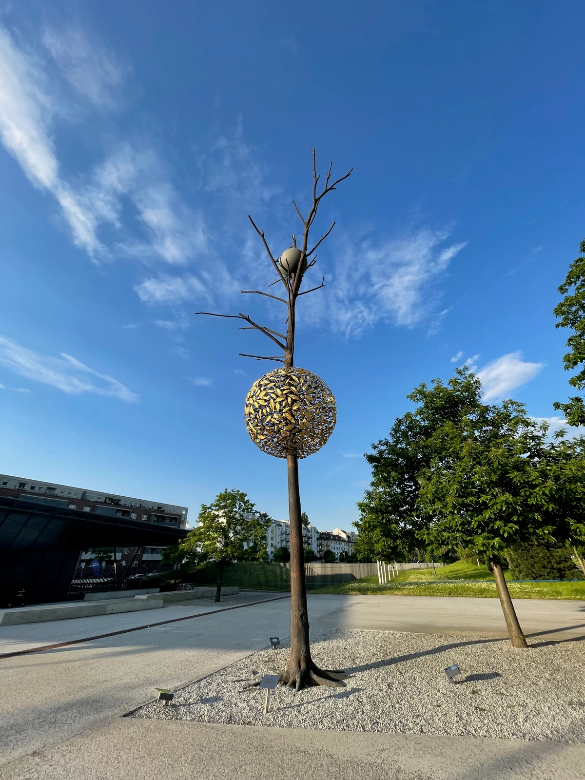 Giuseppe Penone's 'Gravity and Growth' sculpture, a tree-like structure with a spherical, golden leaf-like element and a grey sphere at the top, set against a blue sky with clouds.