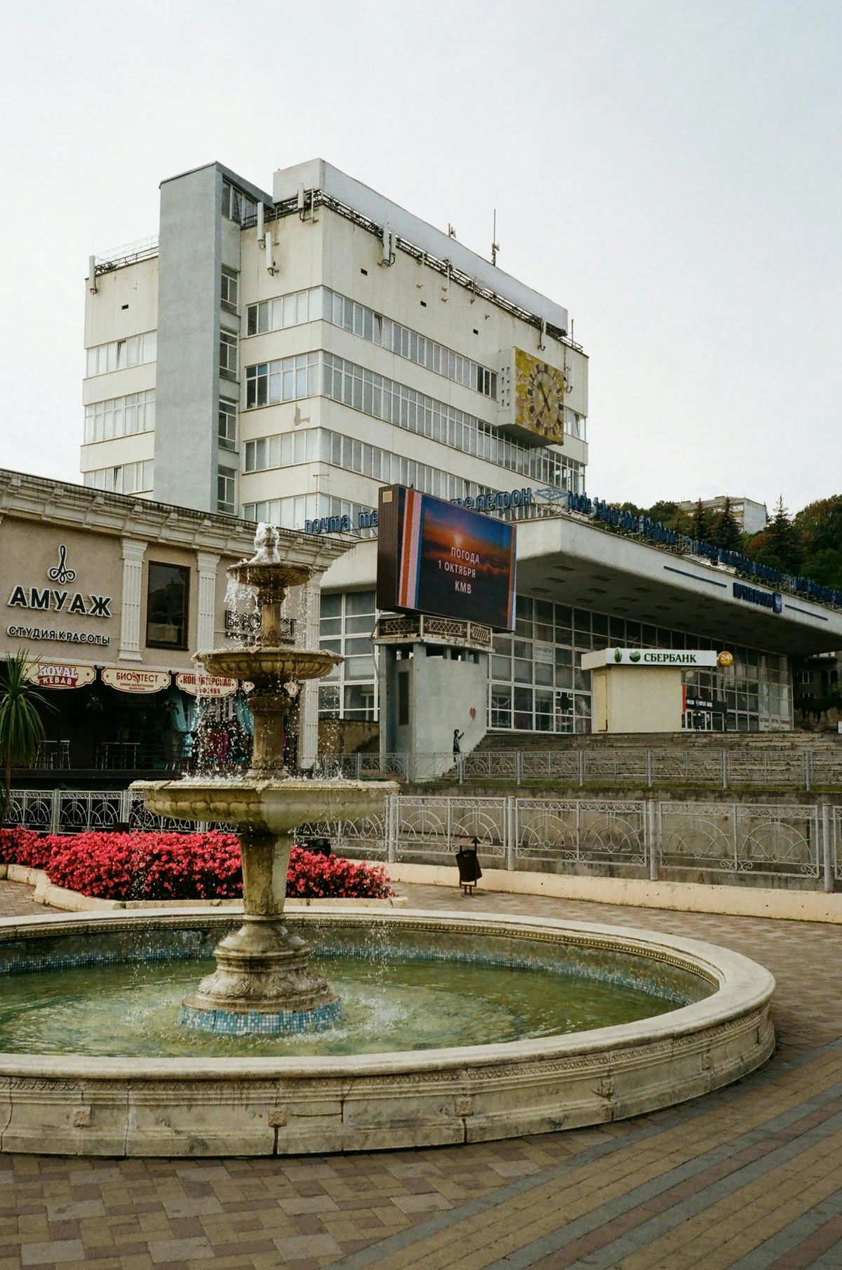 A historic ornate fountain in a bustling European town square, surrounded by white mid-century buildings, cobblestone streets, and vibrant red flowerbeds, symbolizing civic pride and community heritage.