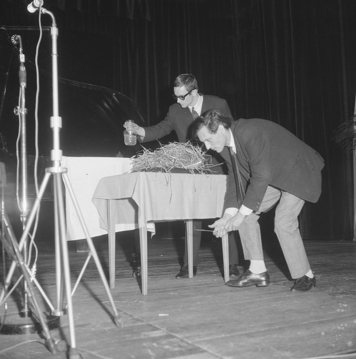 Two men in suits performing with a nest and a jar of liquid on a table, possibly a Fluxus event.