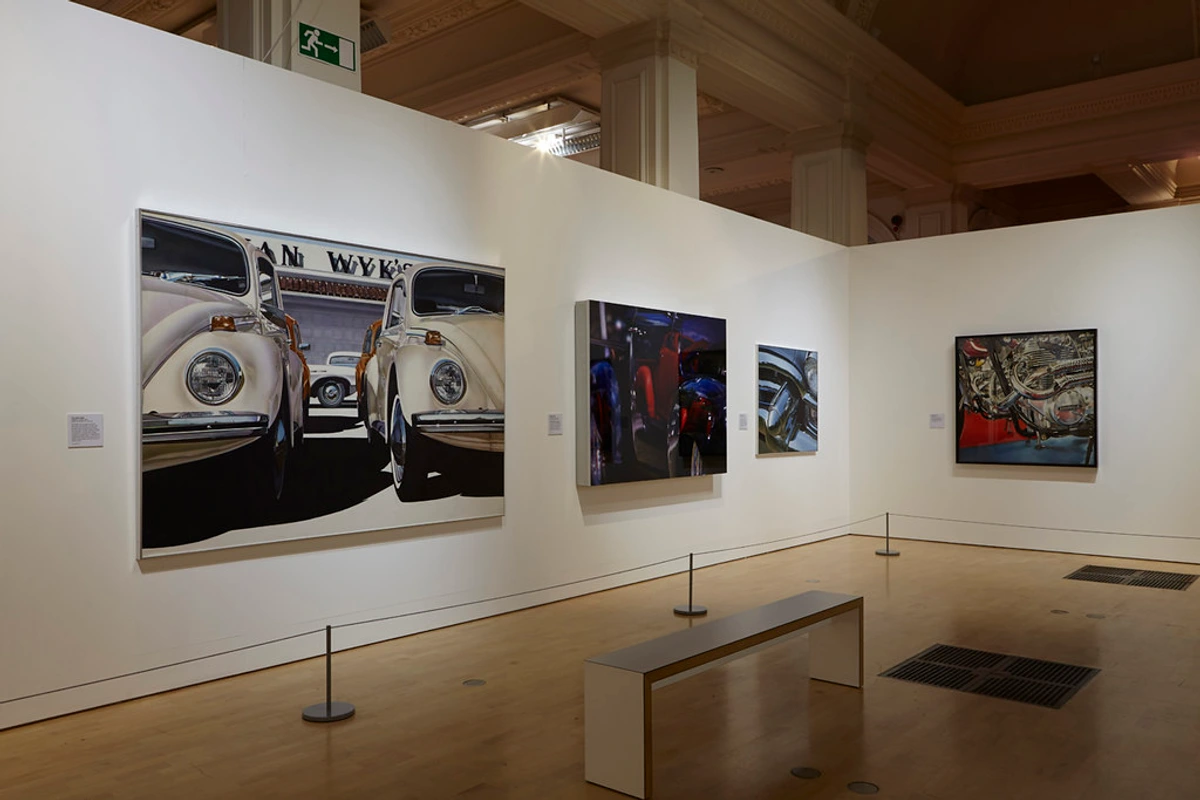 Hyperrealistic paintings of VW Beetles and a car engine displayed in an art gallery.