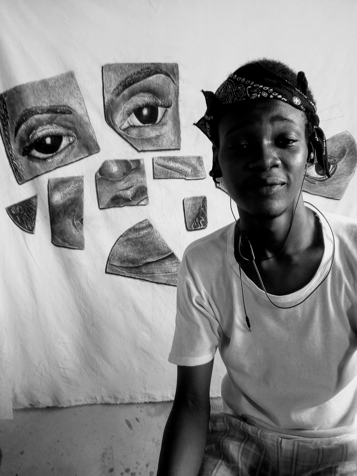 Aliu Kabiru Olatunji, a young man wearing a bandana and headphones, posing in front of his hyperrealistic charcoal drawings of facial features.