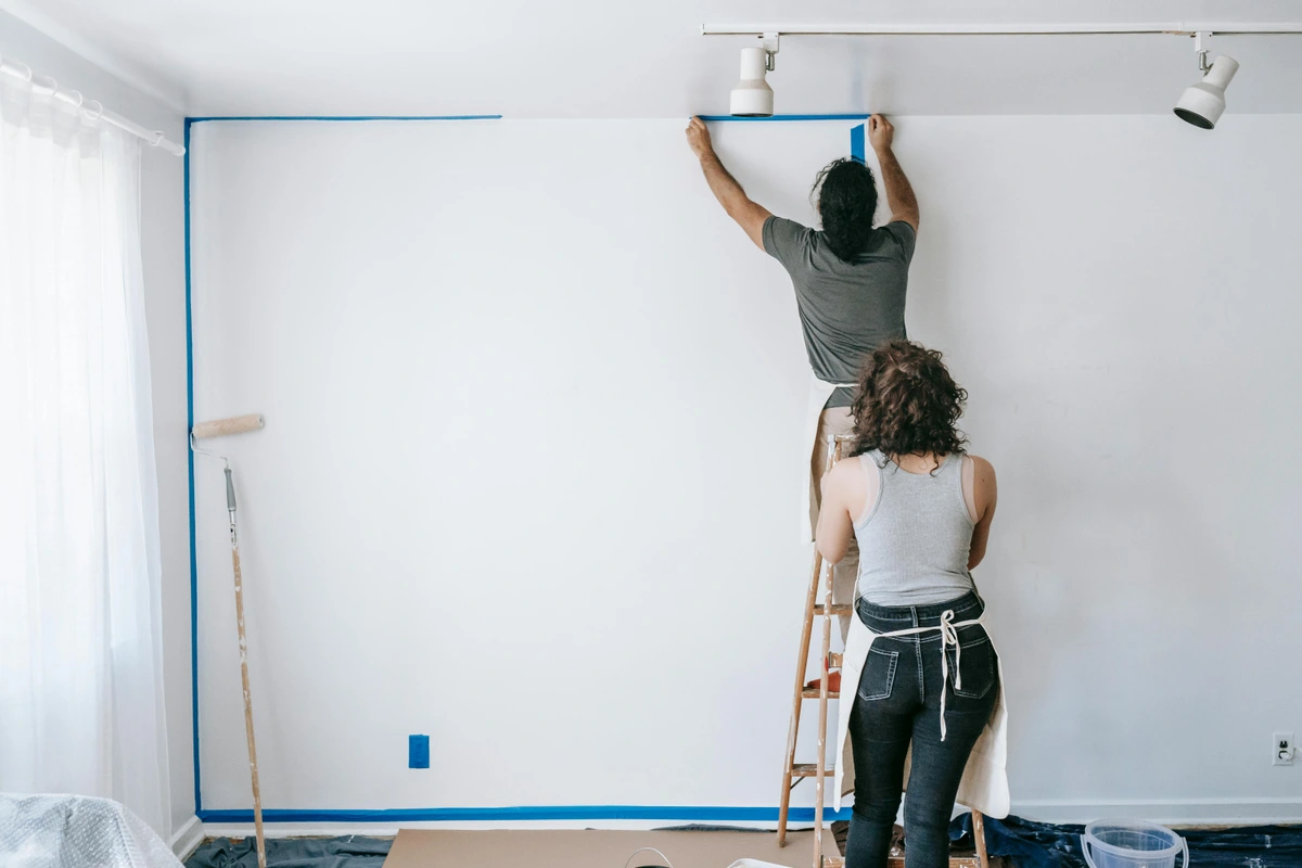 Man applying blue painter's tape to a wall using a ladder for art or painting projects, DIY home improvement setup with tools and protective cloths laid on the floor.