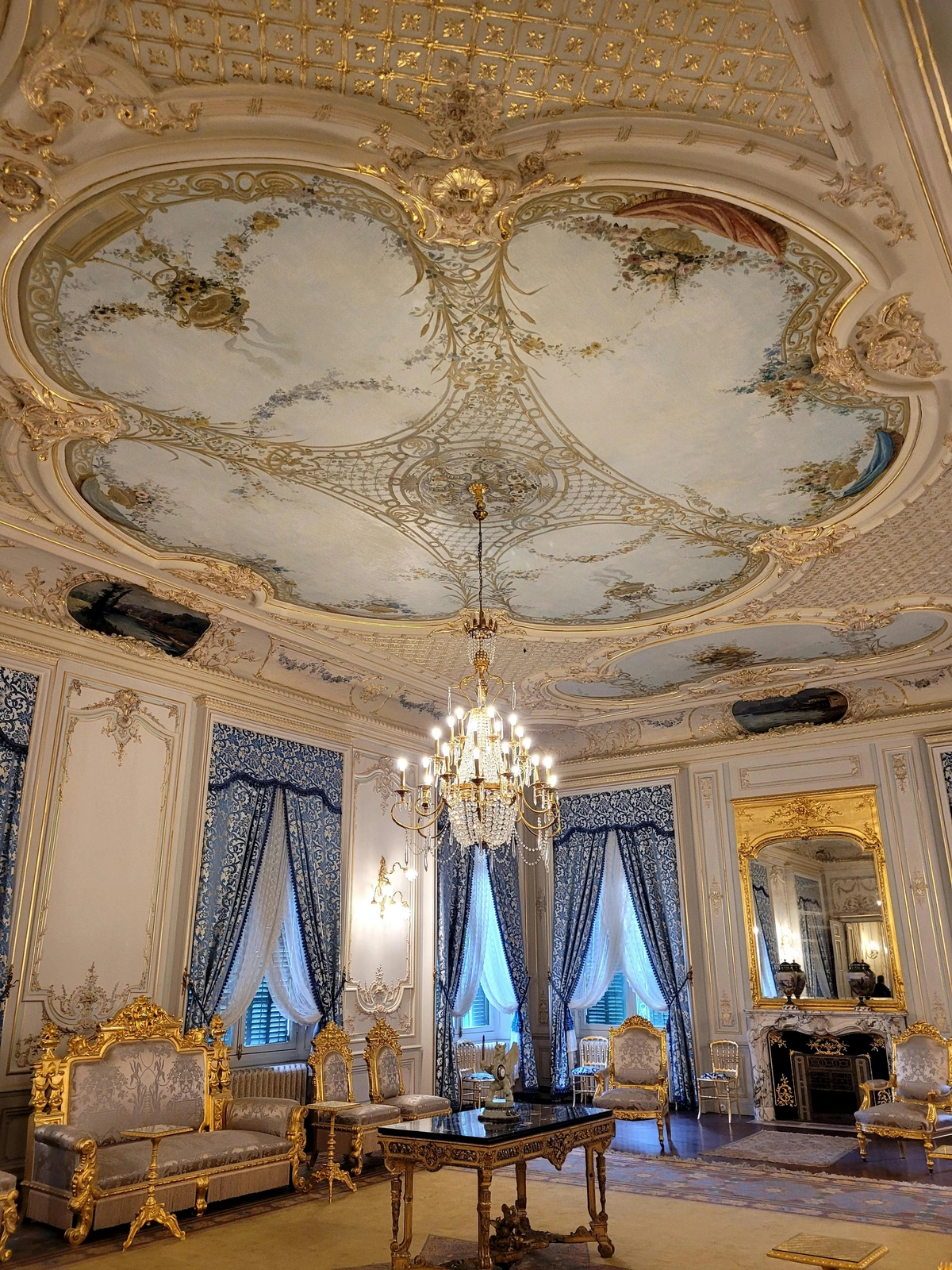 Ornate French Rococo interior featuring a grand gilded chandelier, intricate ceiling murals, luxurious furniture, and blue damask curtains.