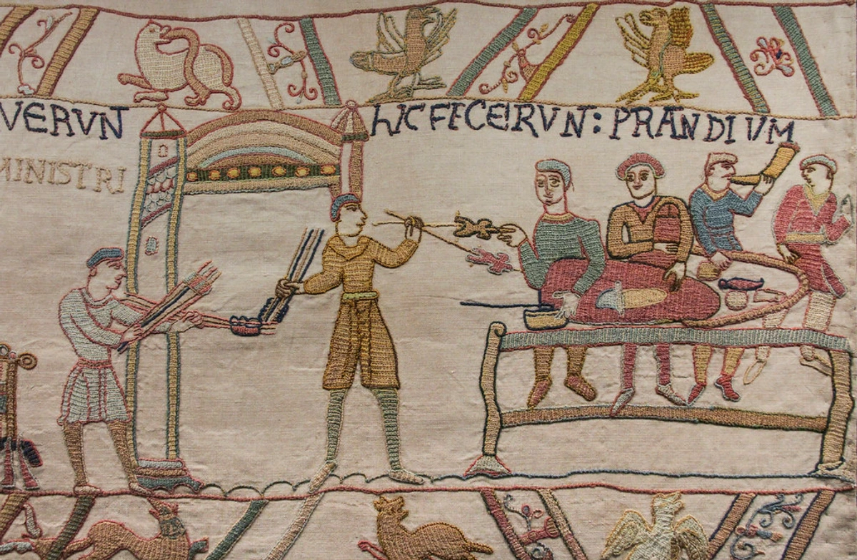 Bayeux Tapestry scene depicting figures in medieval attire, some preparing food and others seated at a table, with Latin inscriptions above.