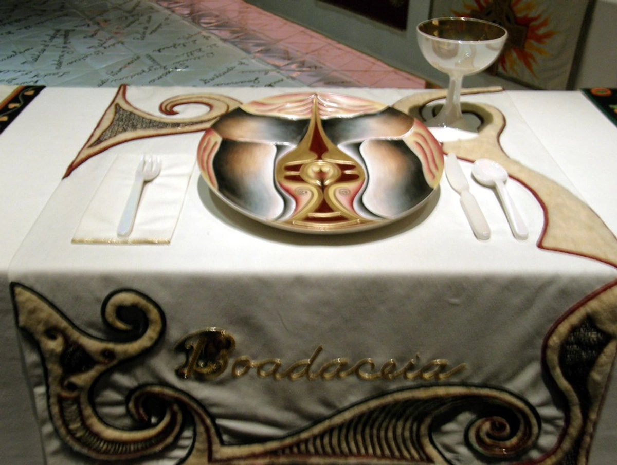 The Dinner Party by Judy Chicago, featuring the Boadaceia place setting with a ceramic plate, goblet, and cutlery on a decorated table runner.