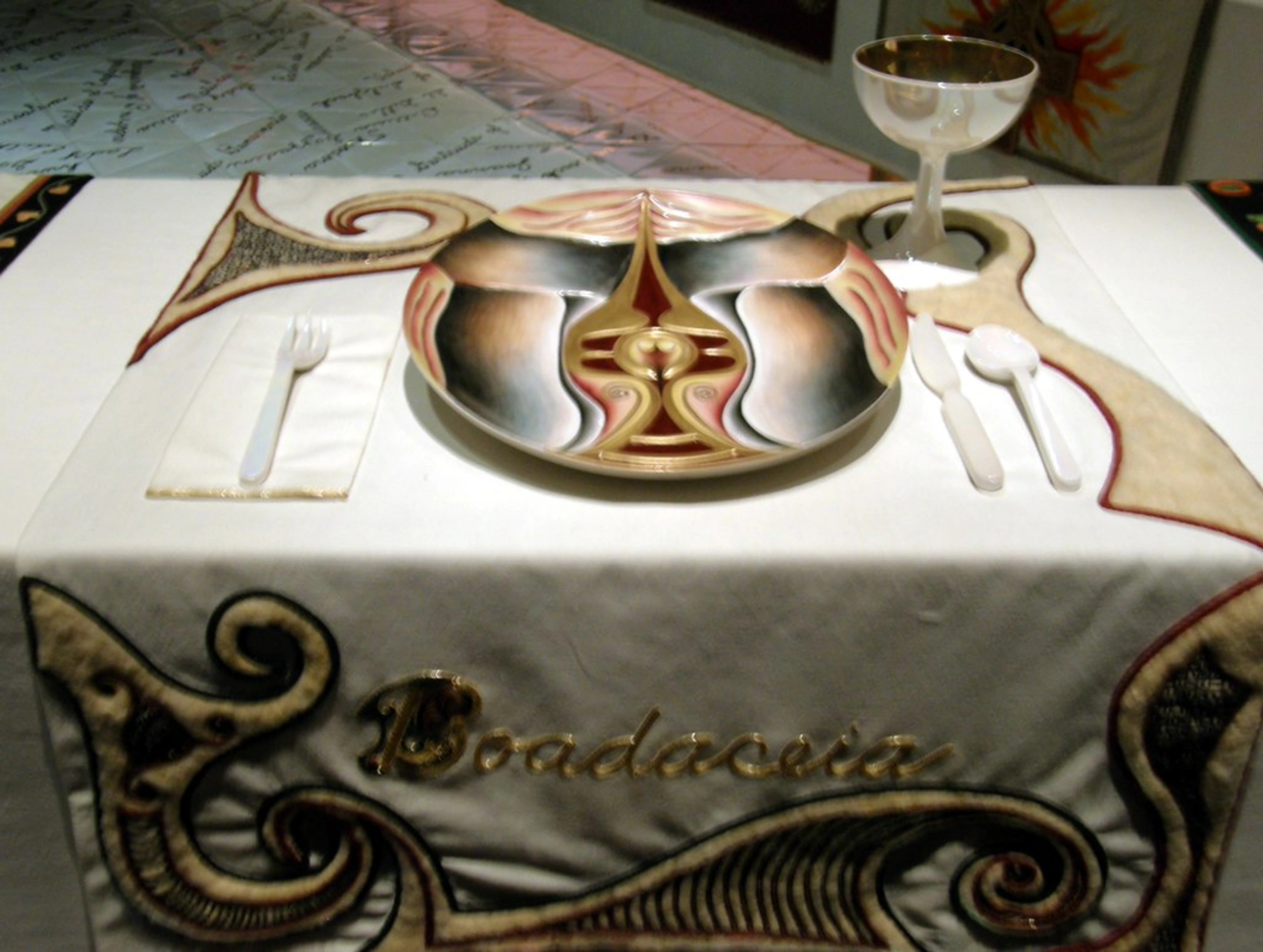 The Dinner Party by Judy Chicago, featuring the Boadaceia place setting with a ceramic plate, goblet, and cutlery on a decorated table runner.