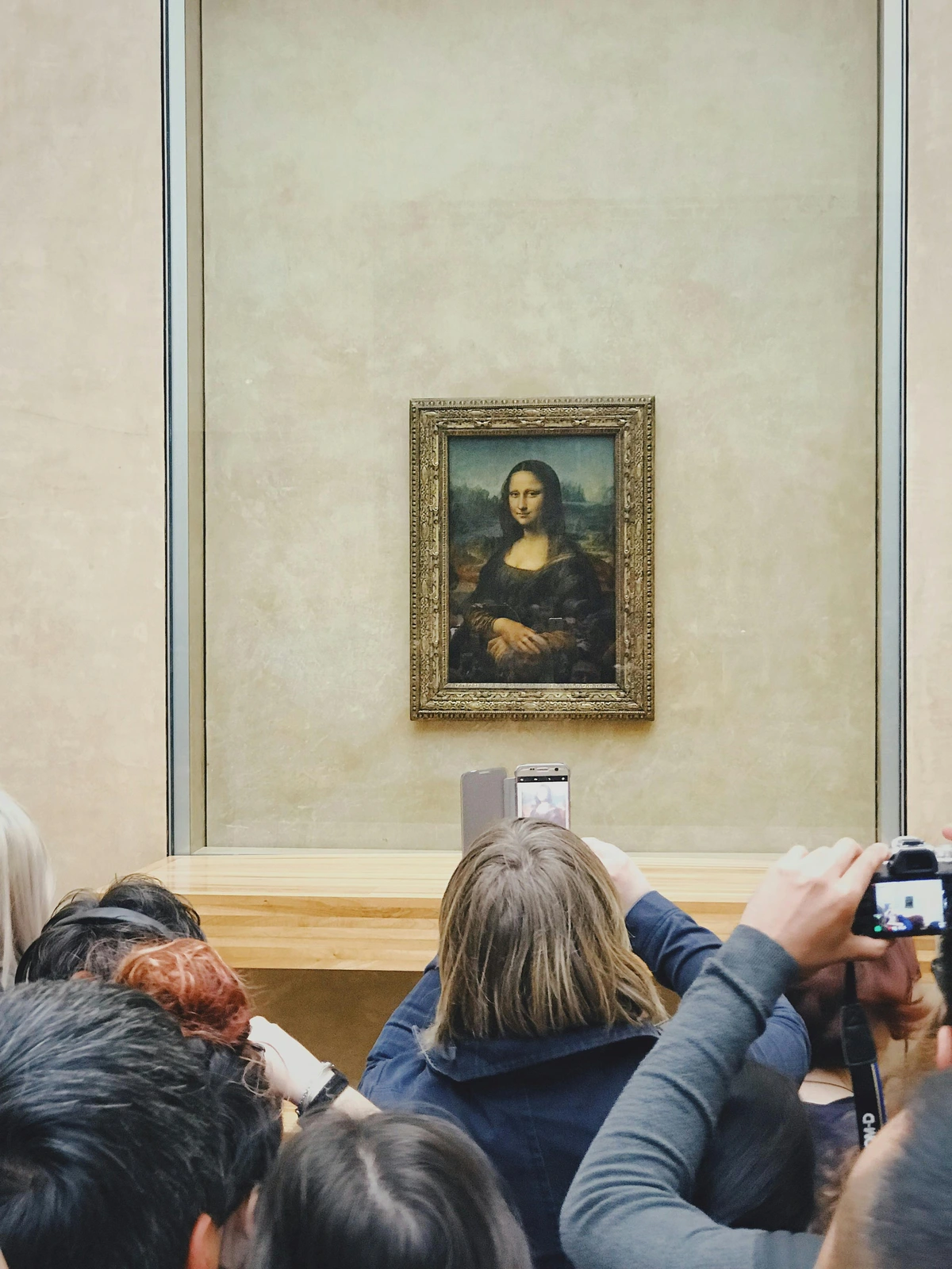Mona Lisa portrait with people taking photographs in a museum setting, showcasing the iconic Renaissance artwork in a framed glass display.