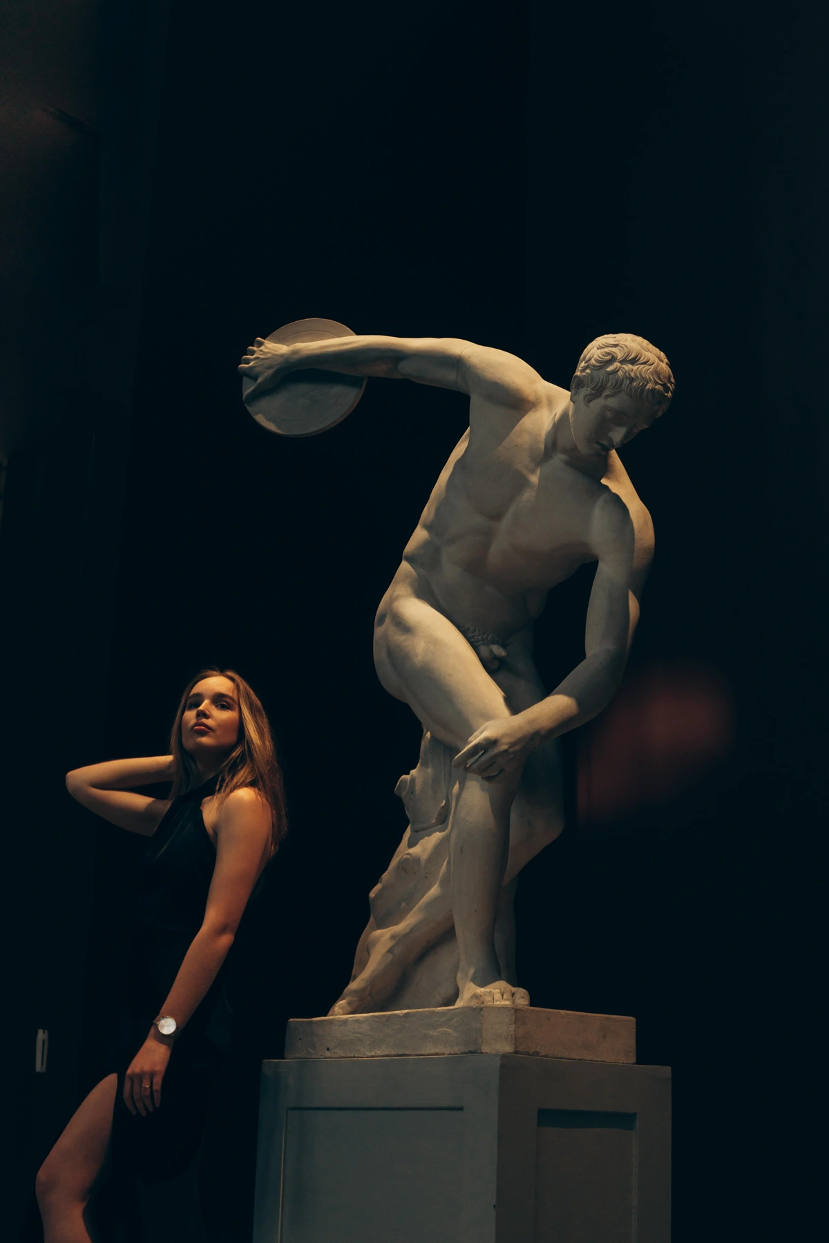 A woman in a black dress poses next to a marble statue of the Discus Thrower in a dimly lit museum.