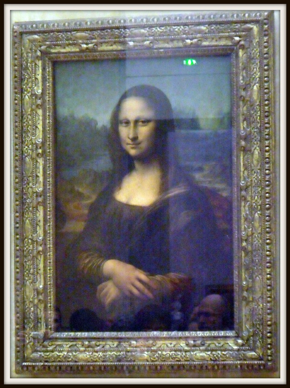 The Mona Lisa painting by Leonardo da Vinci, displayed in an ornate gilded frame behind protective glass.