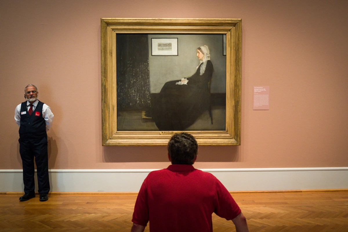 A museum guard stands to the left of James McNeill Whistler's famous painting 'Whistler's Mother', while a visitor in a red shirt views the artwork from behind.