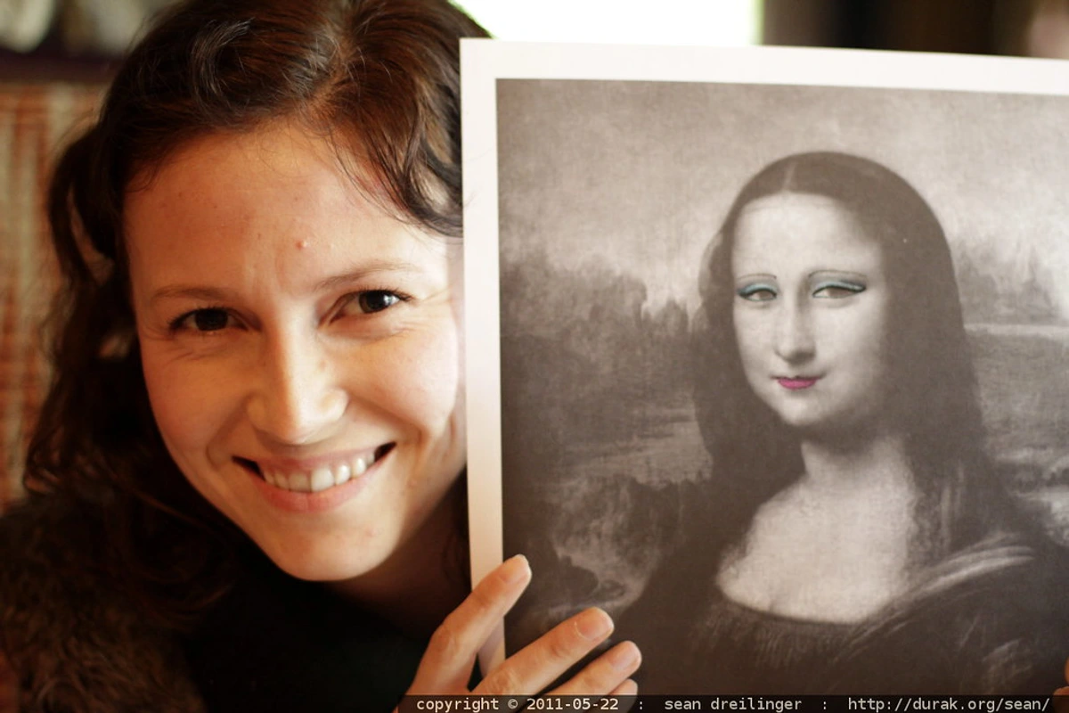 Woman holding a print of the Mona Lisa with modern, colorful makeup applied to her face.