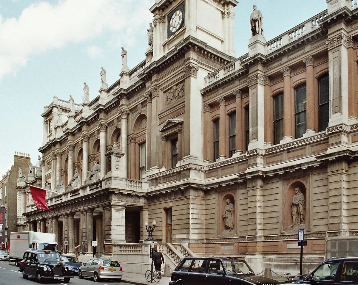 The Royal Academy of Arts building in London, a grand Neoclassical structure with a prominent clock tower, columns, and statues, viewed from the street with traffic.
