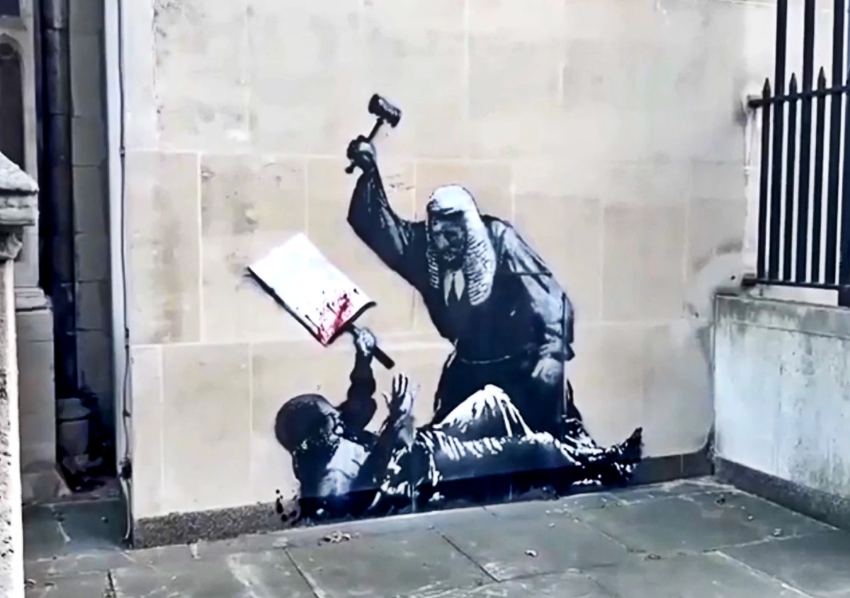 Banksy mural depicting a judge in traditional robes smashing a book with a gavel, while a person lies on the ground.