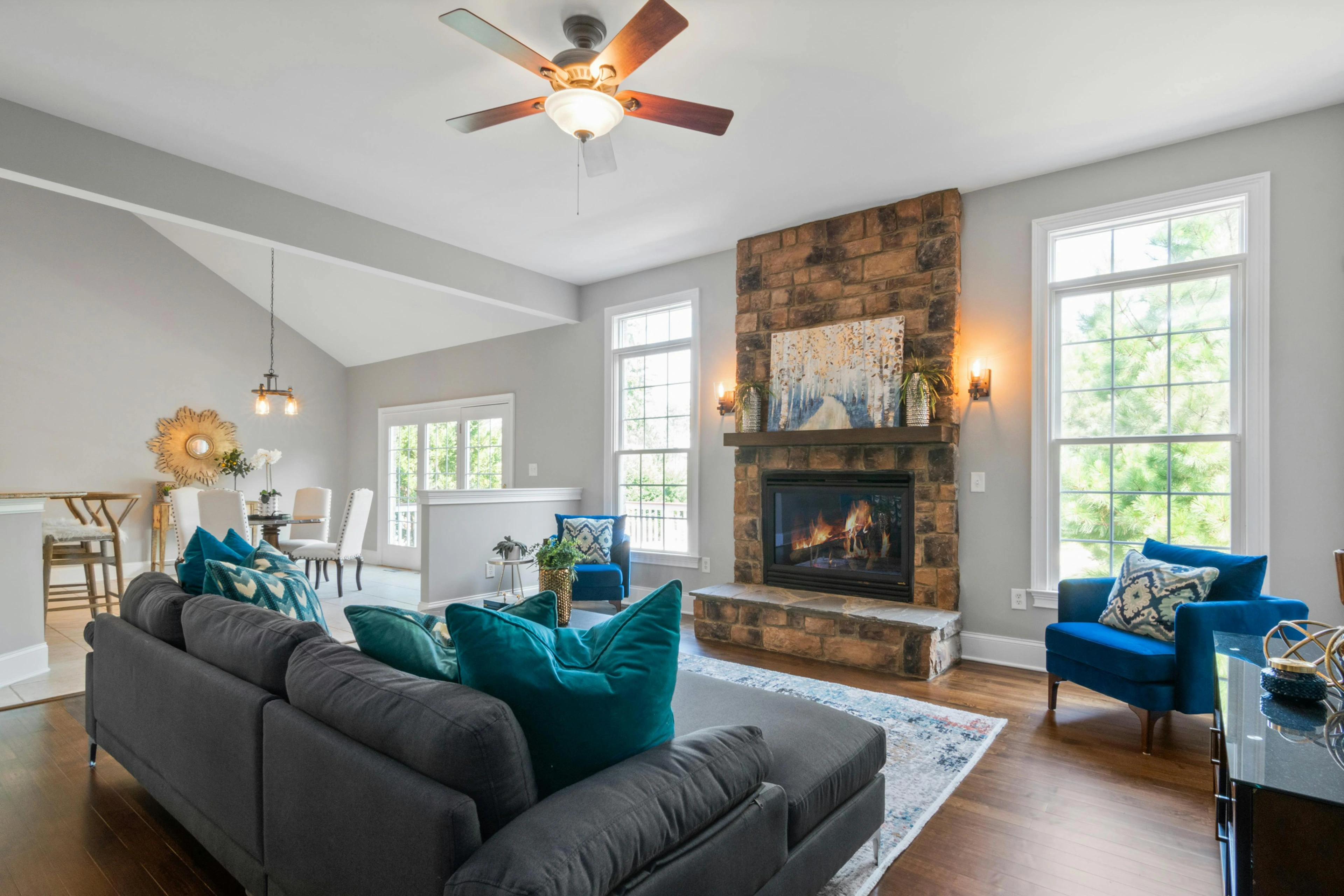 Modern living room with a dark gray sectional sofa adorned with vibrant teal and patterned blue pillows, facing a rustic stone fireplace with a colorful abstract painting above it. A plush blue armchair sits to the right, complementing the decor.