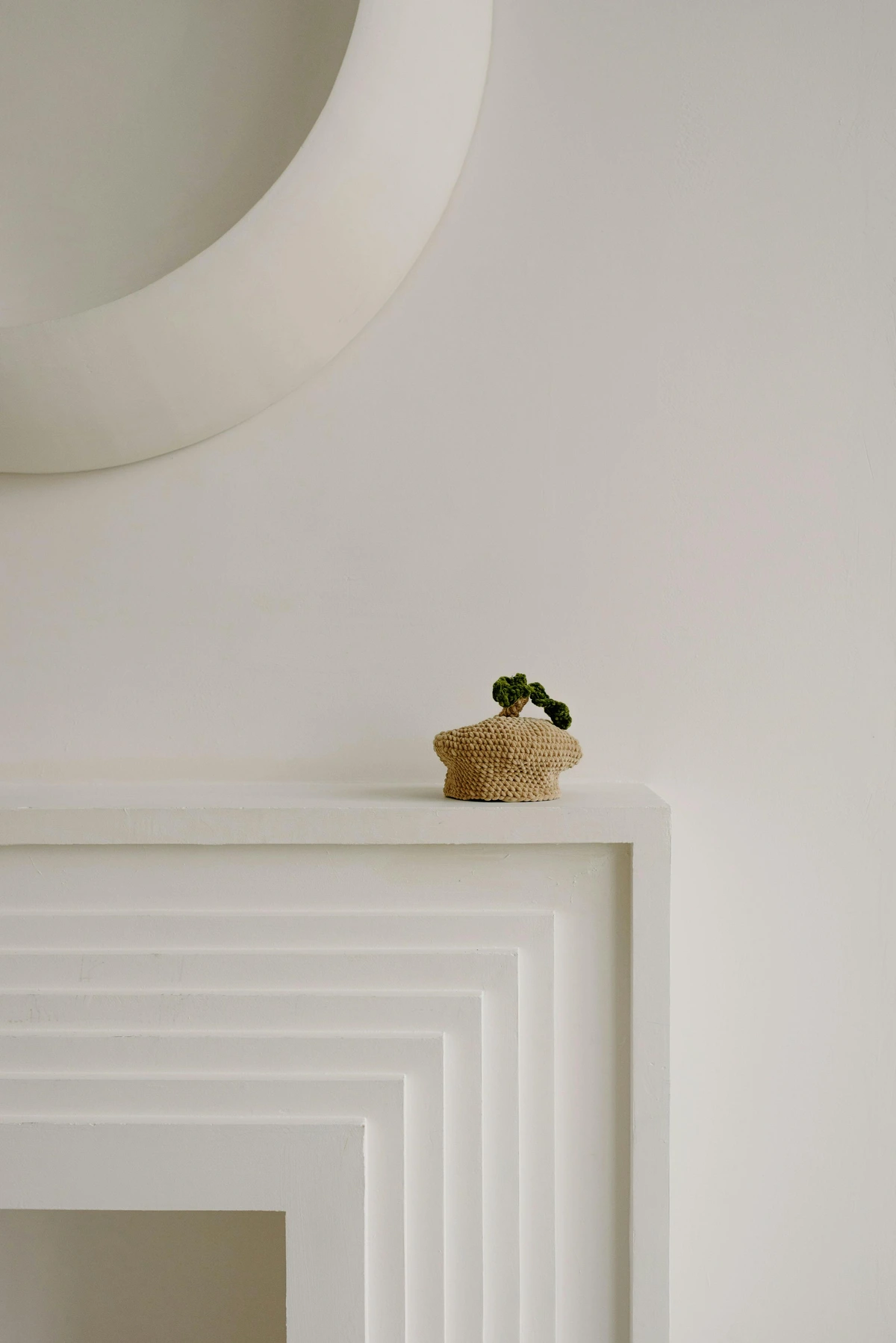A small, crocheted decorative item resembling a plant in a pot sits on a modern, layered fireplace mantel.