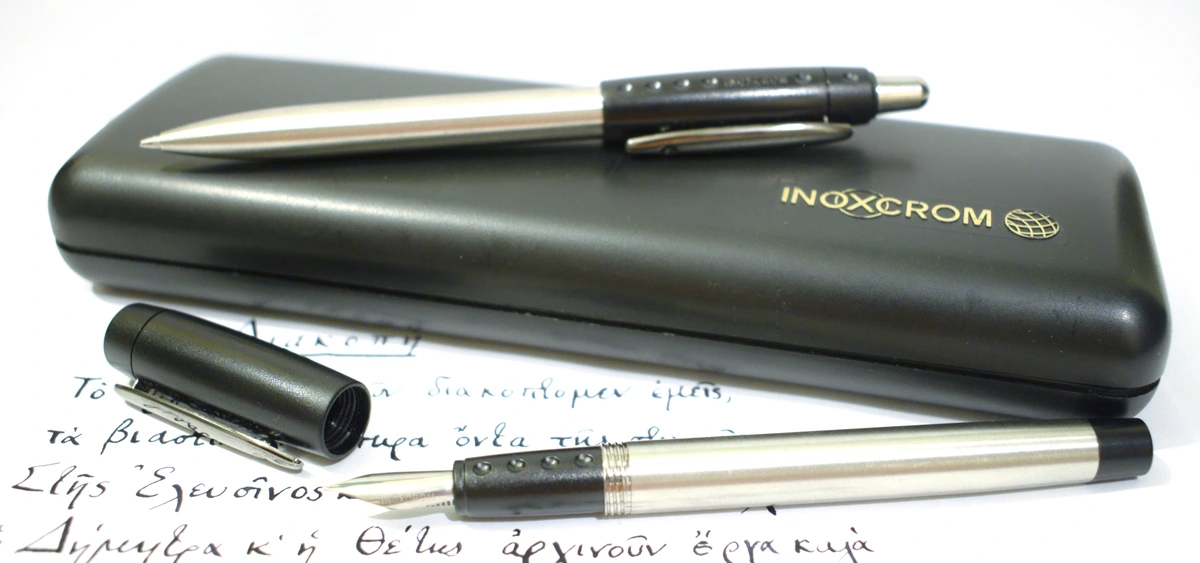 A traditional fountain pen and ballpoint pen rest on paper with Greek script, illustrating pen hold for calligraphy.