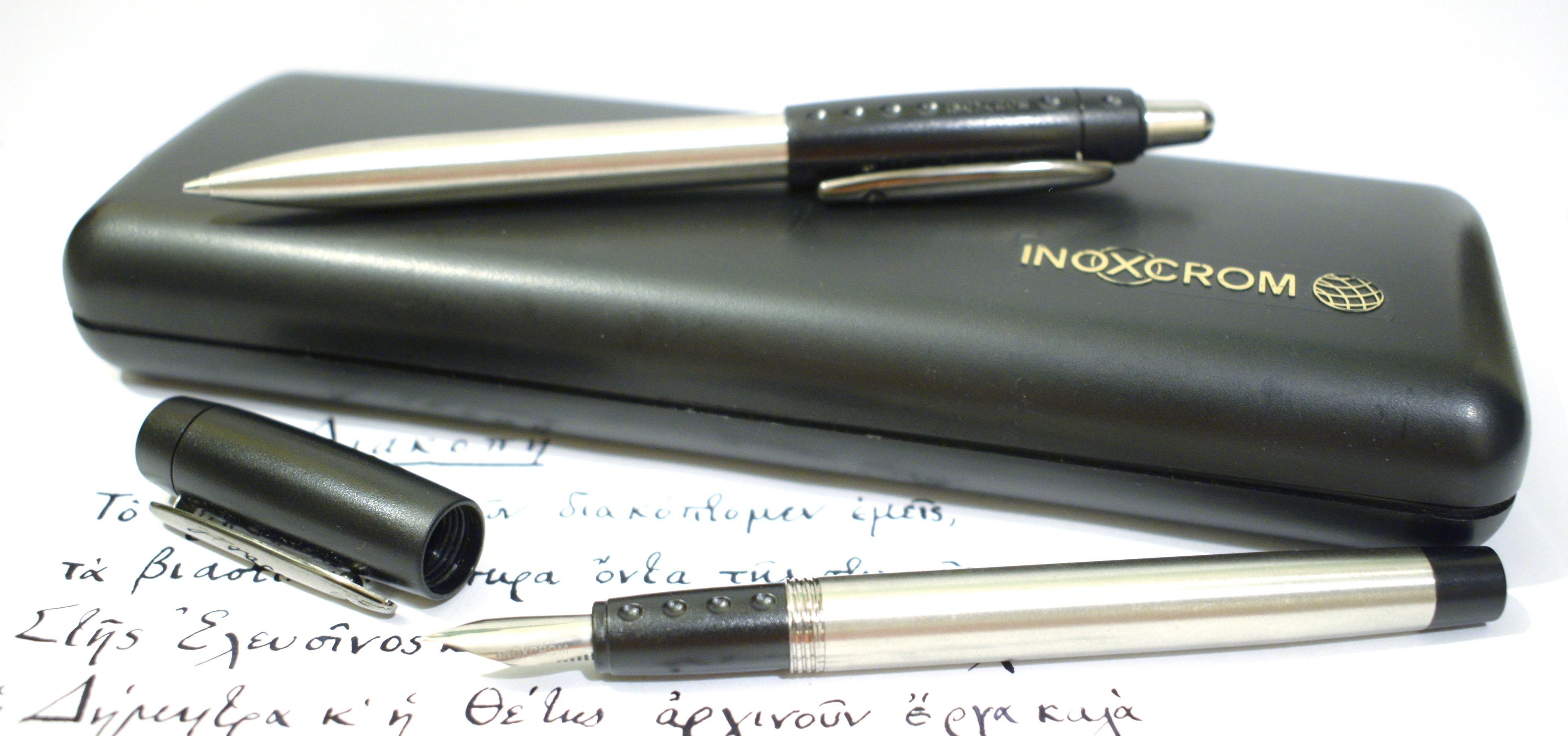 A black Inoxcrom pen case with a silver fountain pen and a silver ballpoint pen on top, resting on a piece of paper with handwritten Greek text.