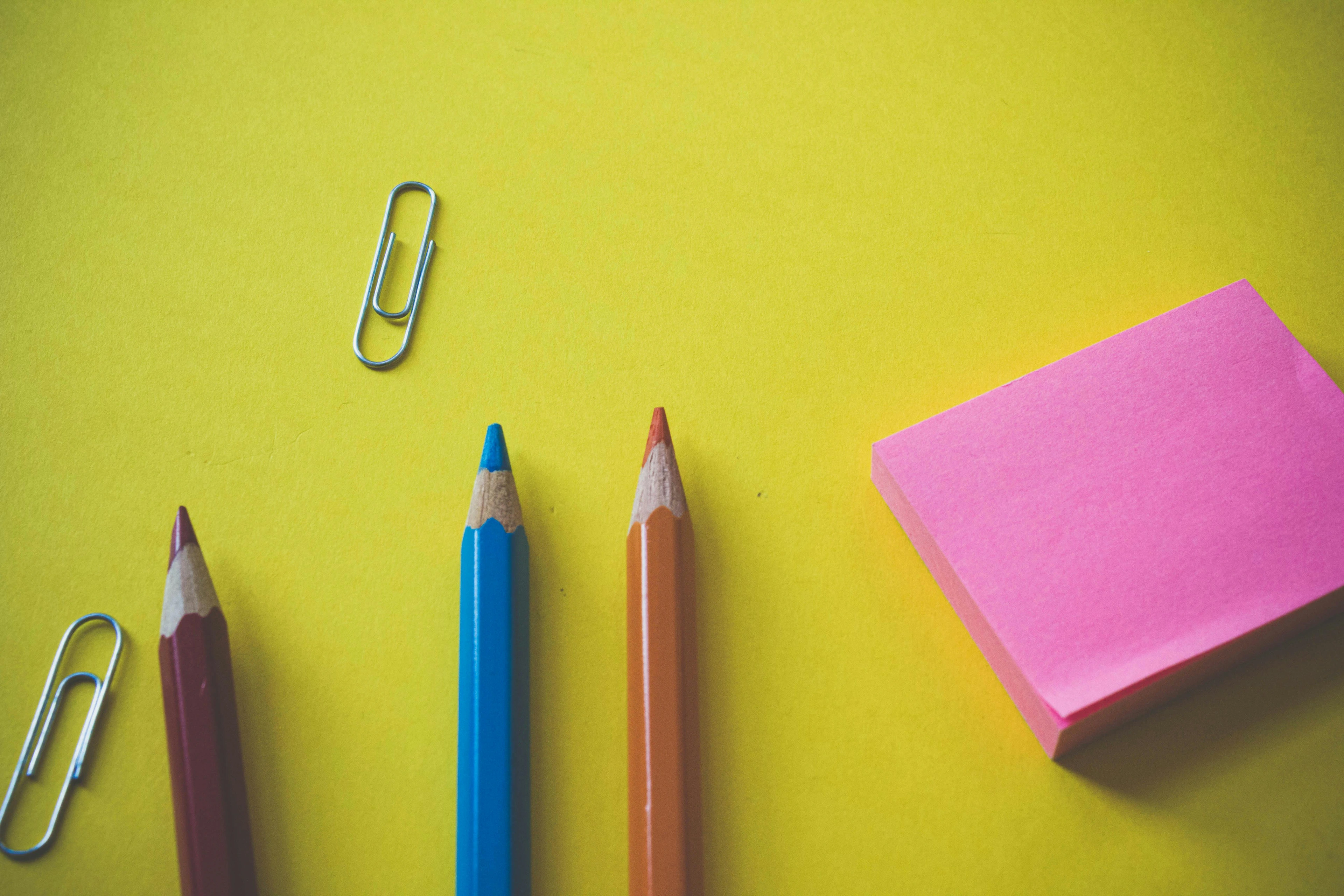 Top view of colorful pencils, paper clips, and a pink sticky note pad on a yellow background, representing writing materials.
