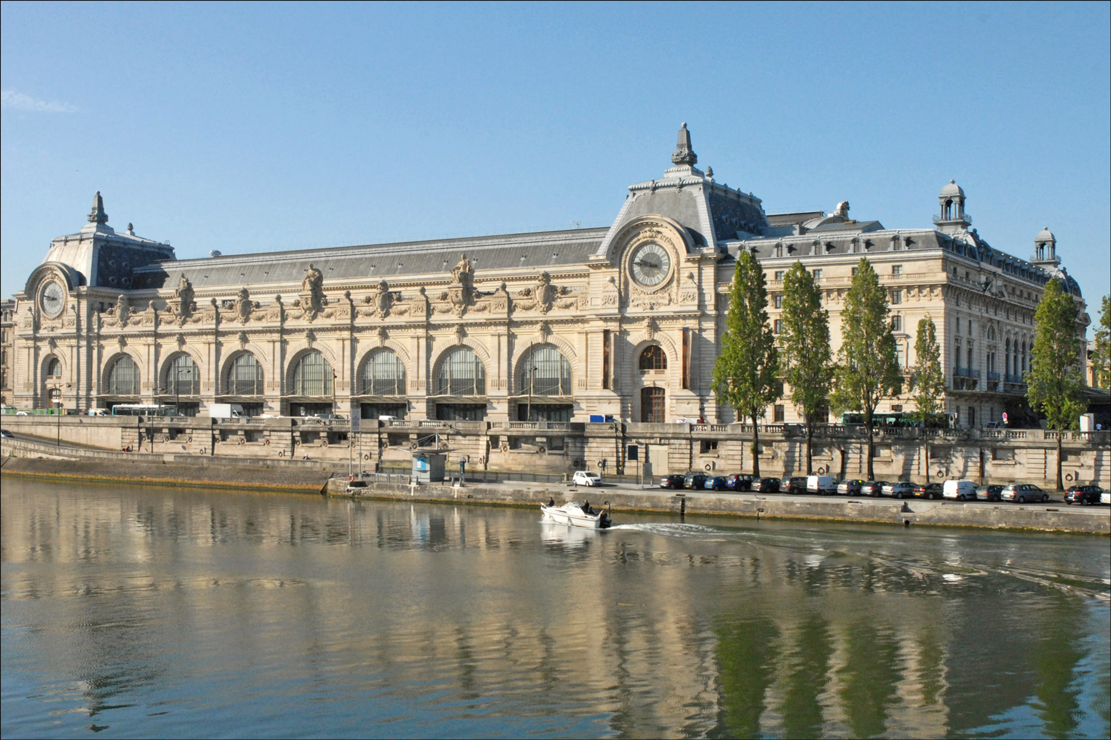 The Musée d'Orsay in Paris, a former railway station, viewed from across the Seine River on a sunny day, with a boat on the water.