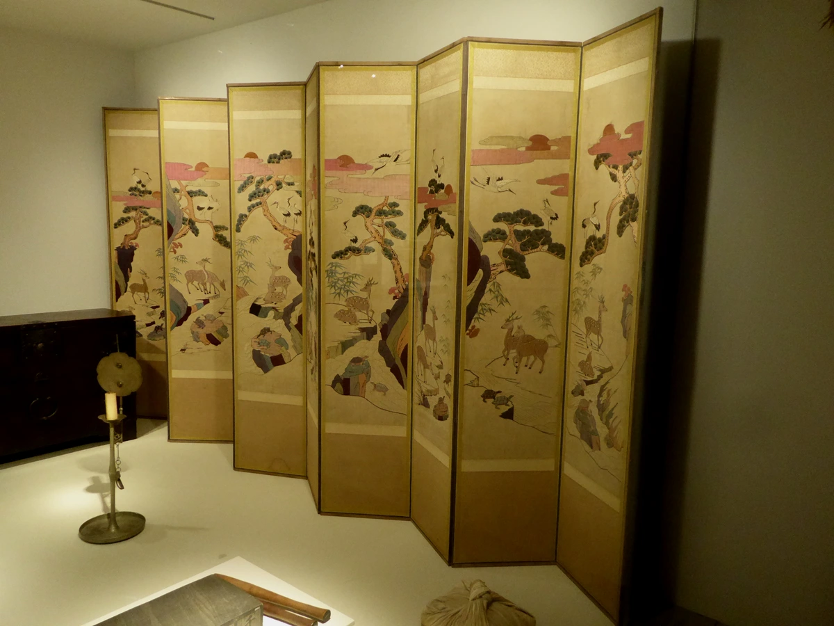 Een geborduurd vouwscherm met kraanvogels, herten en pijnbomen, tentoongesteld in een museum.
