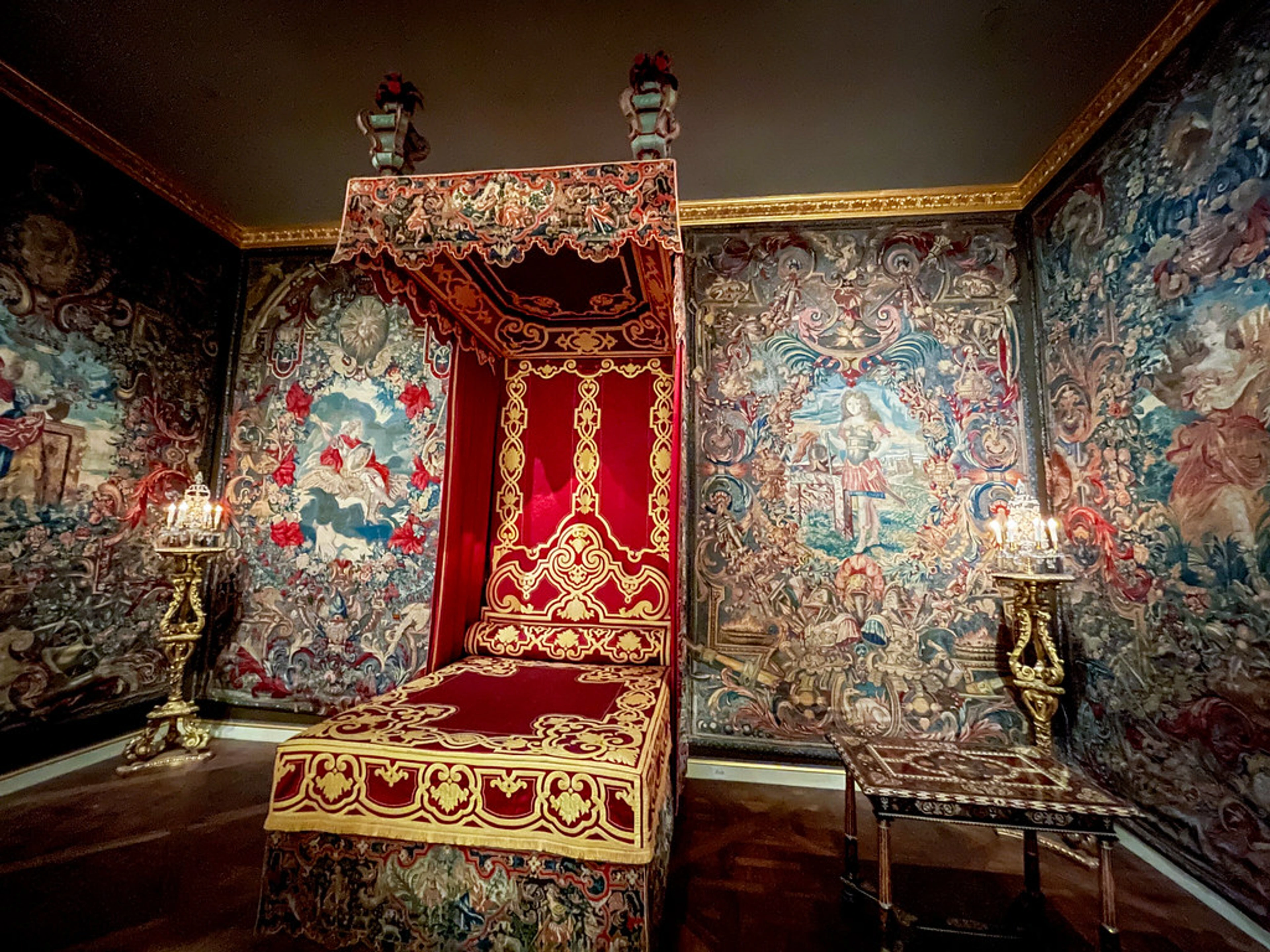 A luxurious bedroom with ornate red and gold canopy bed, surrounded by large, detailed tapestries depicting historical scenes and figures, in the style of Louis XIV decorative arts.