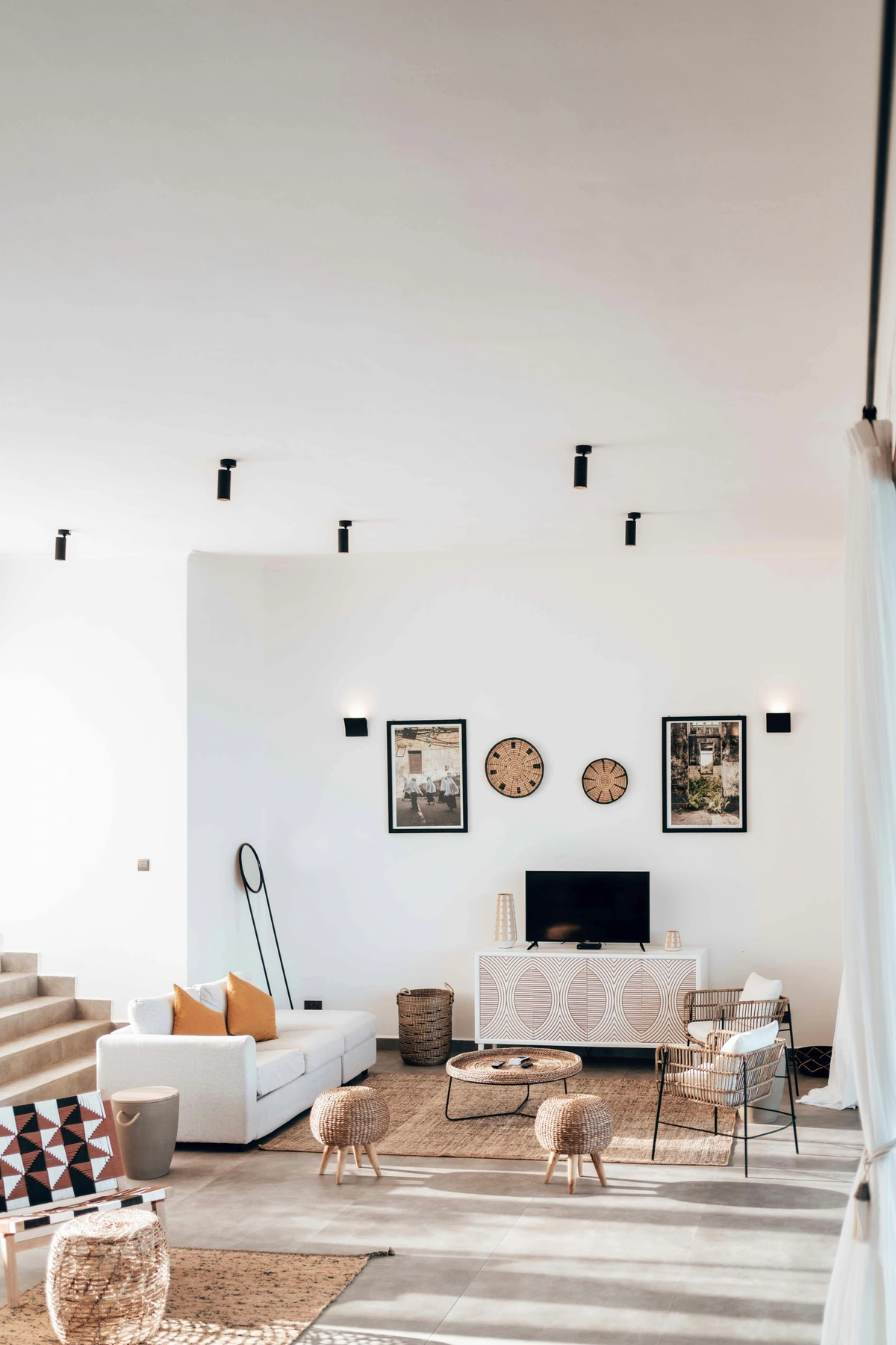 Vertical shot of a bright white living room featuring a white sofa with orange cushions, a round woven coffee table, and braided poufs on a jute rug, showcasing integrated textile art in the interior design.