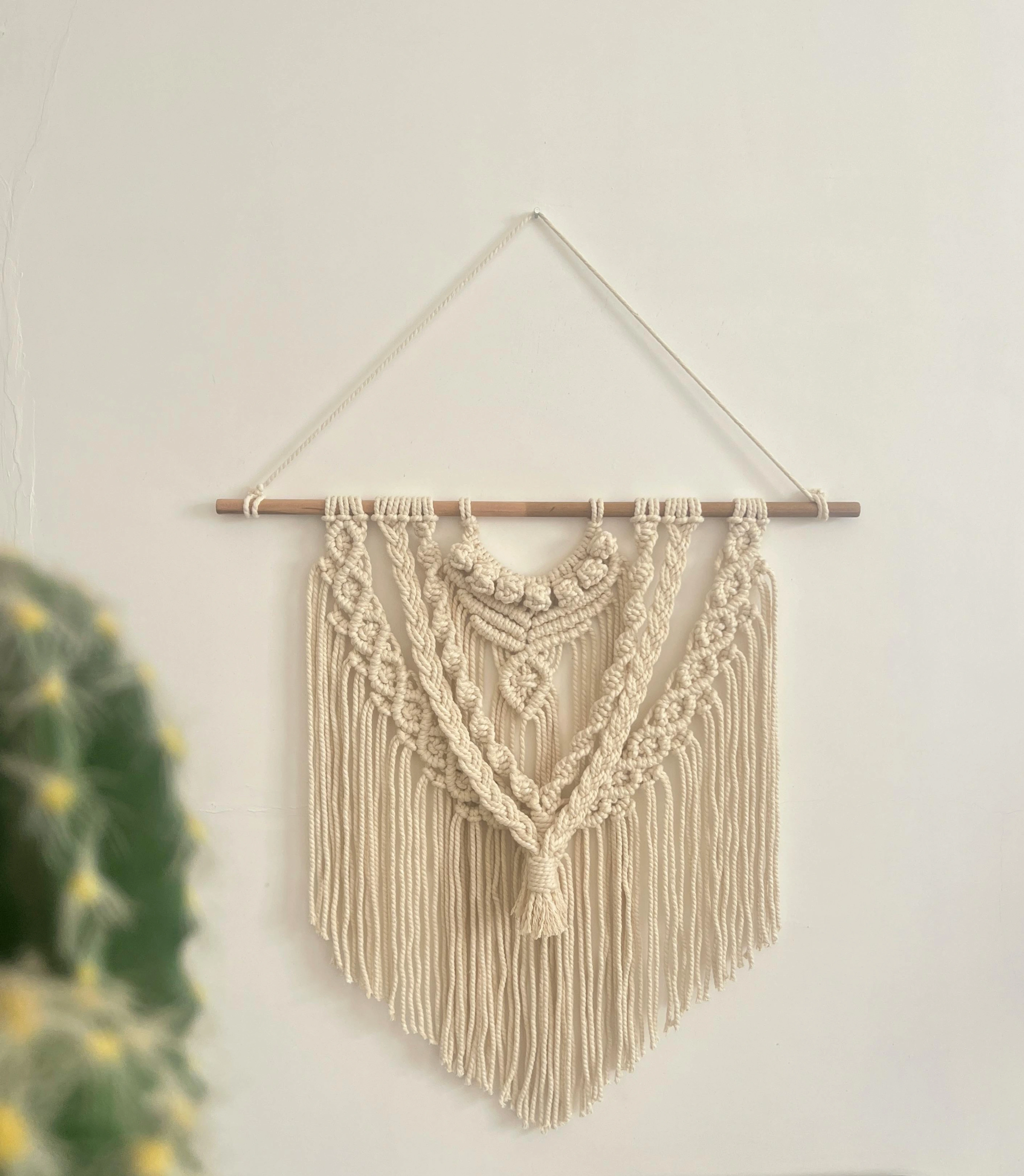 A handmade beige macrame wall hanging with a tassel, suspended from a wooden rod against a white wall, with a blurred cactus in the foreground.