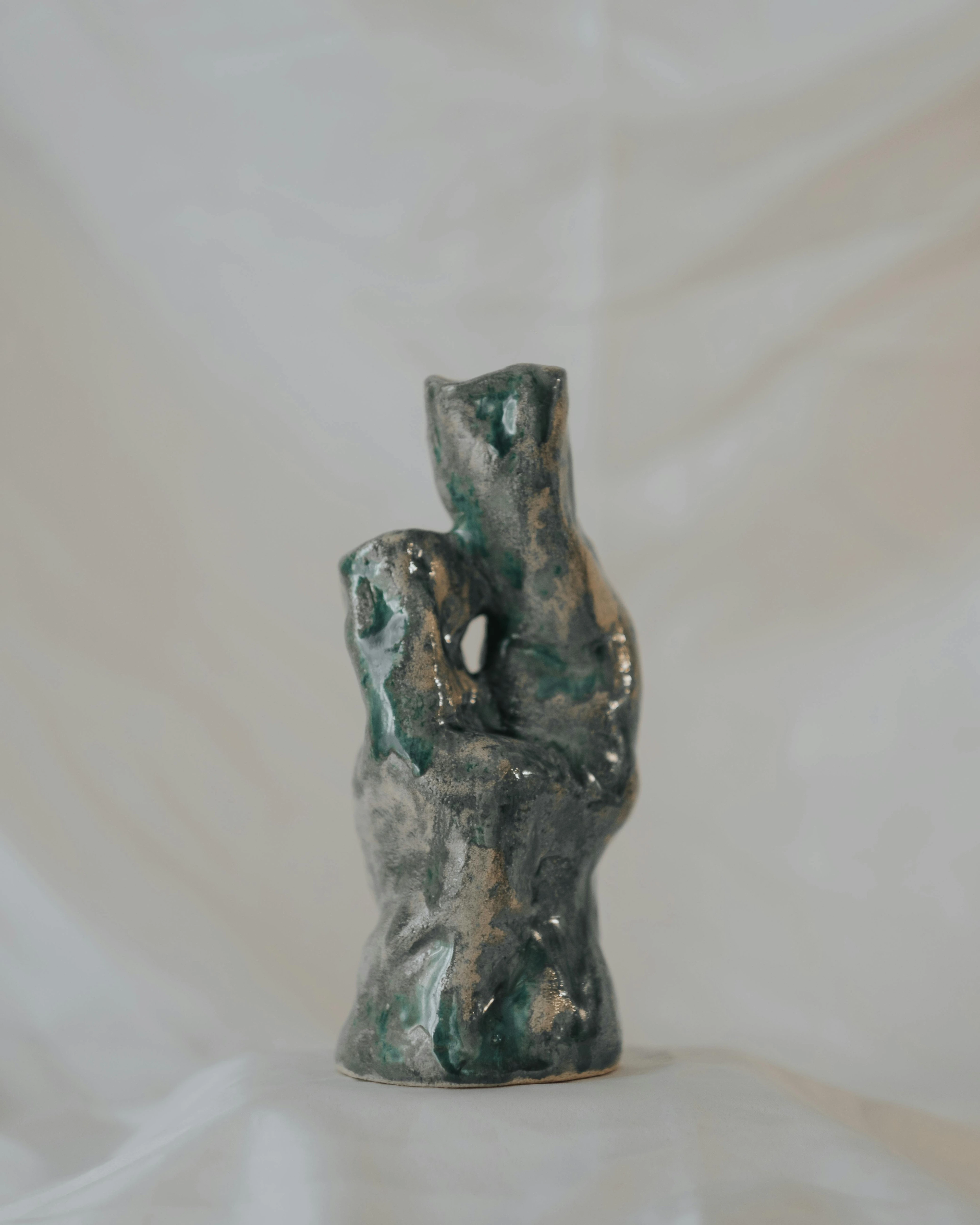 Abstract ceramic sculpture with green and gray glaze, displayed in soft lighting against a white background.