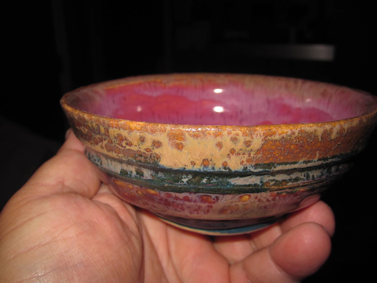 Een hand houdt een kleine, kleurrijke keramische kom met een roze binnenkant en een getextureerde, veelkleurige glazuur aan de buitenkant.