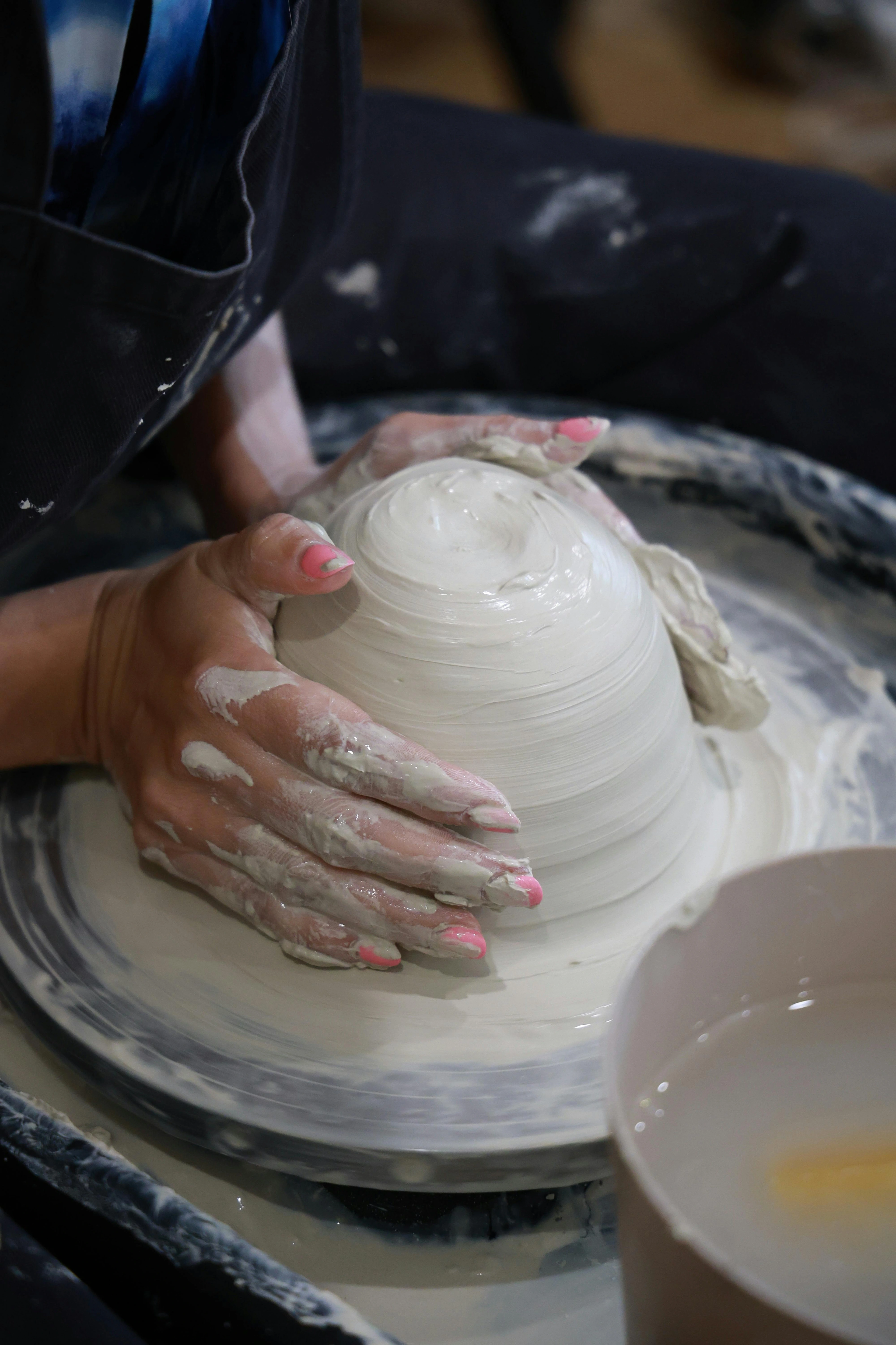 A person's hands with pink-painted nails shaping a wet clay bowl on a pottery wheel, with a bowl of water nearby.