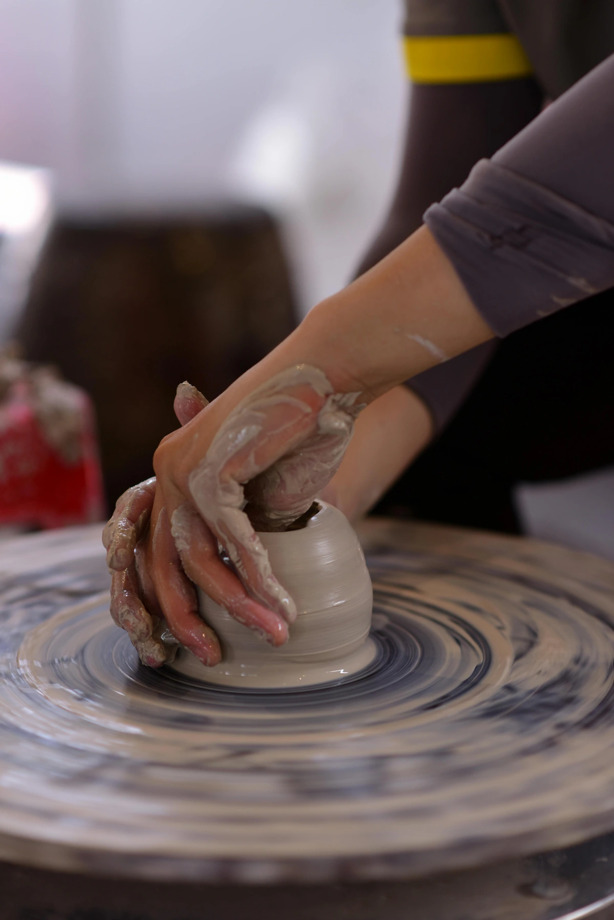 Close-up of hands covered in clay shaping a small pot on a spinning pottery wheel.