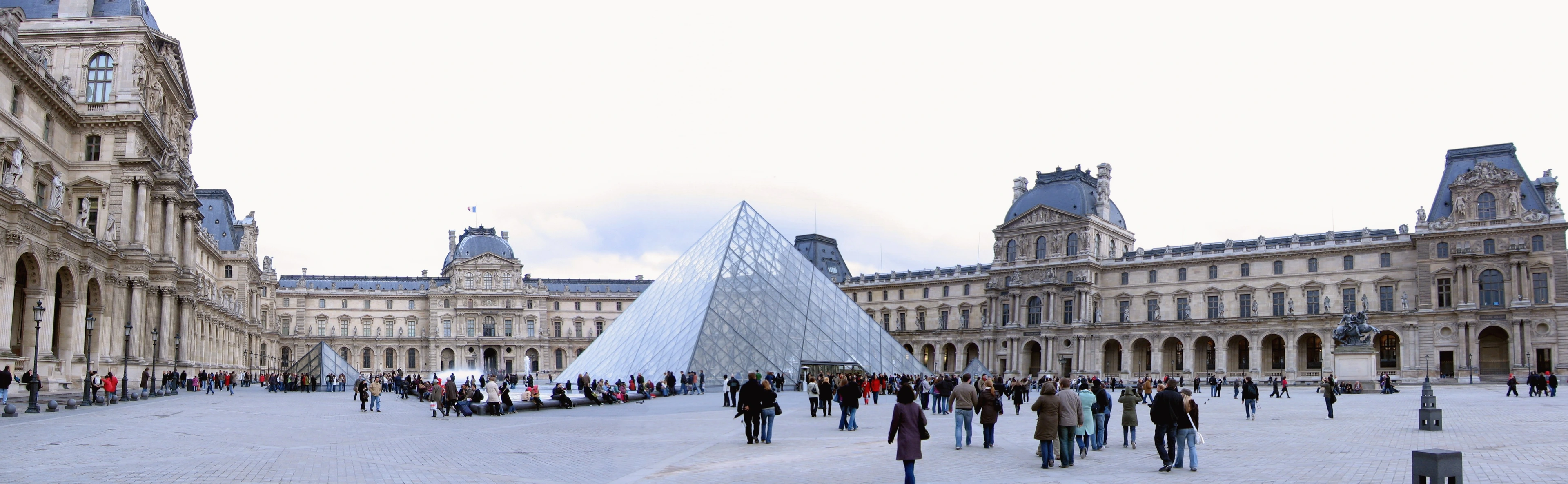Louvre Museum entrance pyramid in Paris, France, with people gathered in the courtyard.