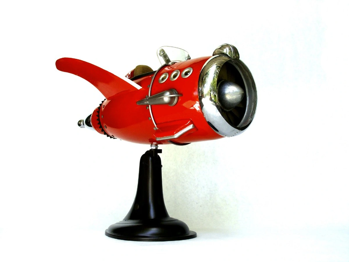 Red found object assemblage sculpture resembling a retro rocket ship, displayed on a black stand.