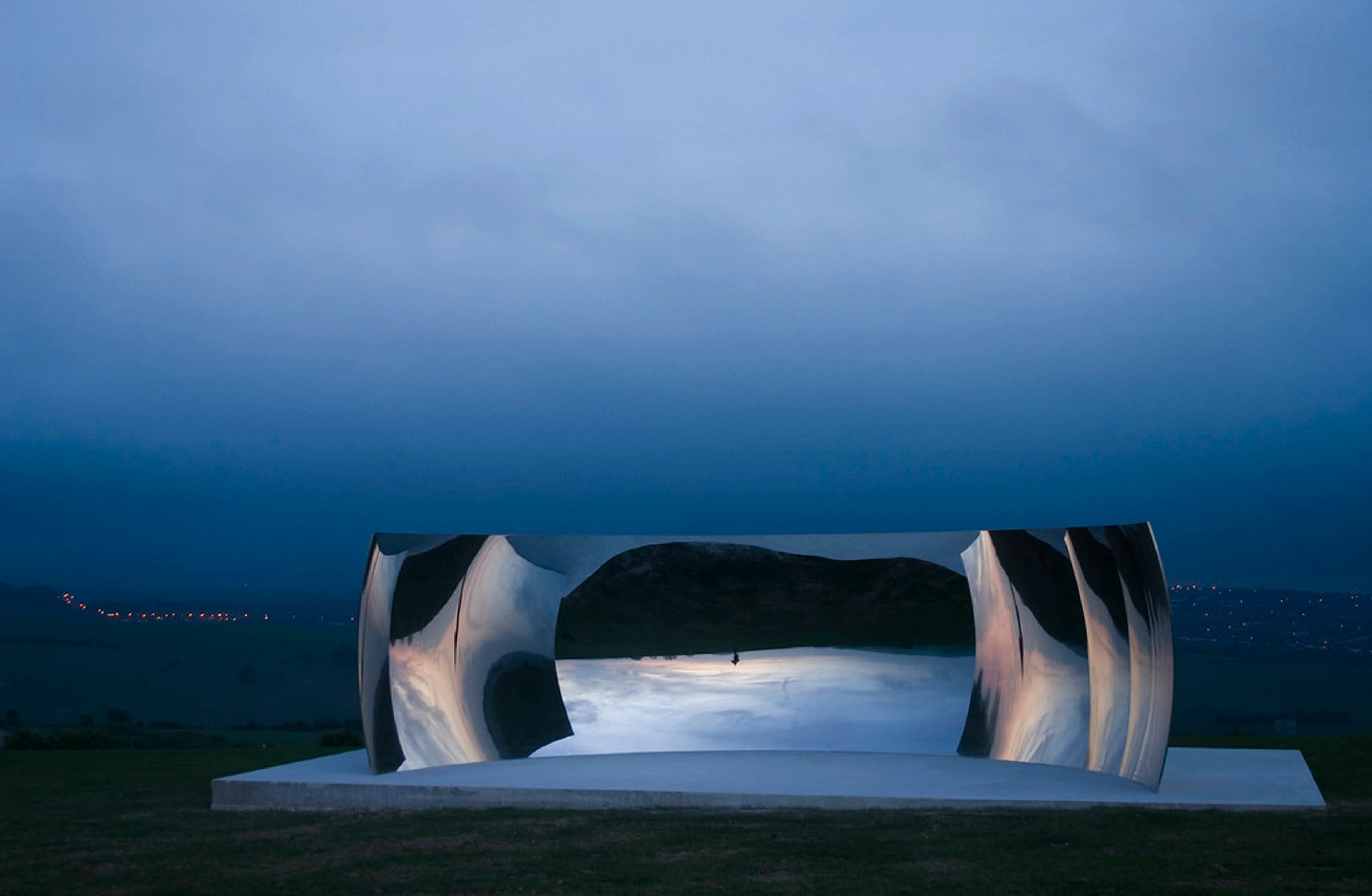 Anish Kapoor's C-Curve (2007) sculpture, a large curved reflective artwork, set outdoors at dusk, reflecting the dark blue sky and distant landscape.