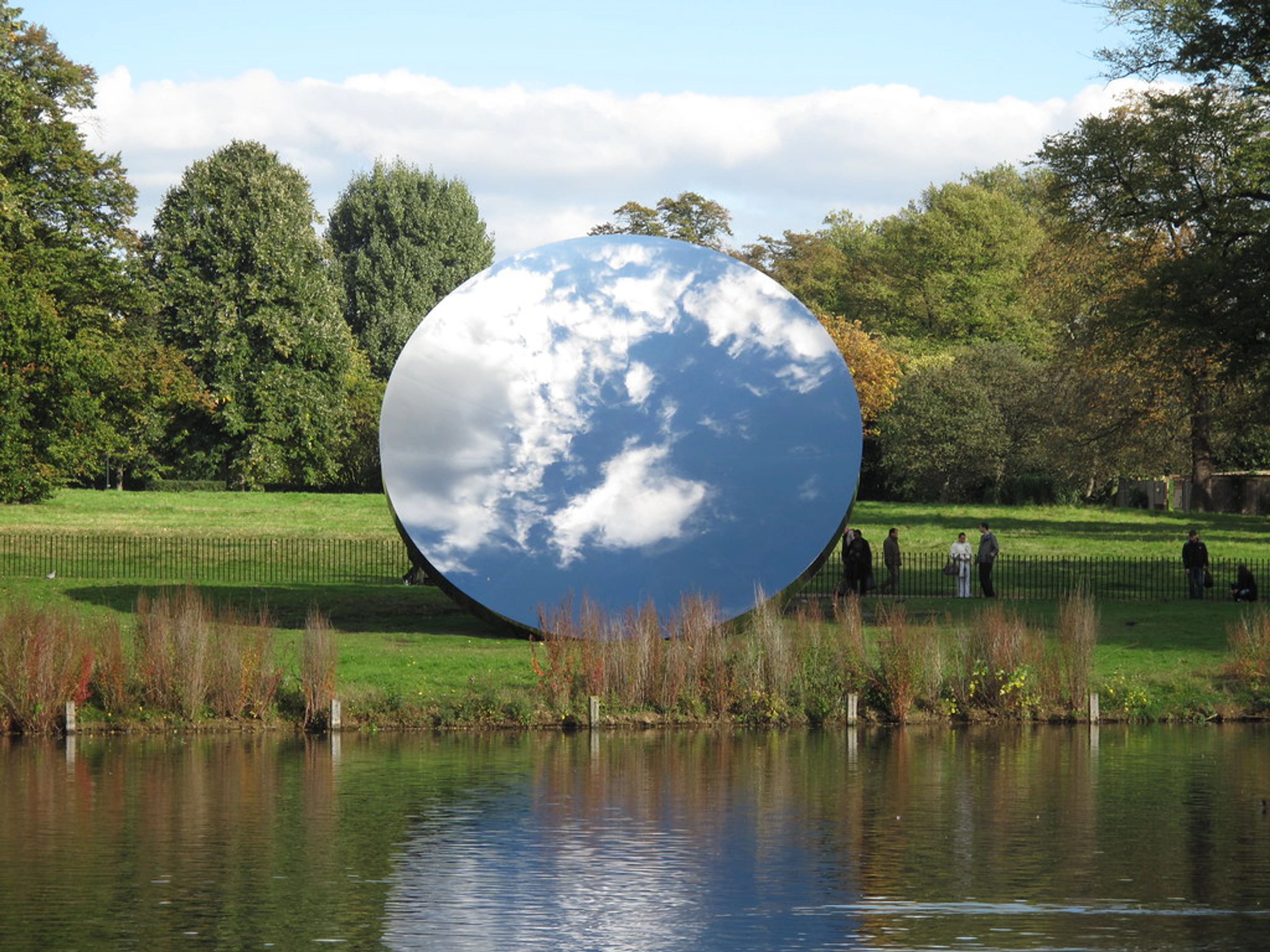 Anish Kapoor's Sky Mirror sculpture reflecting the sky and clouds in Kensington Gardens, London, with trees and a lake.