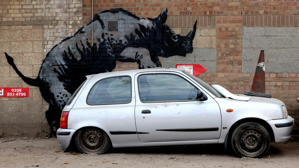 Banksy's 'Rhino' street art on a brick wall with a silver car parked in front. Famous urban graffiti artwork.