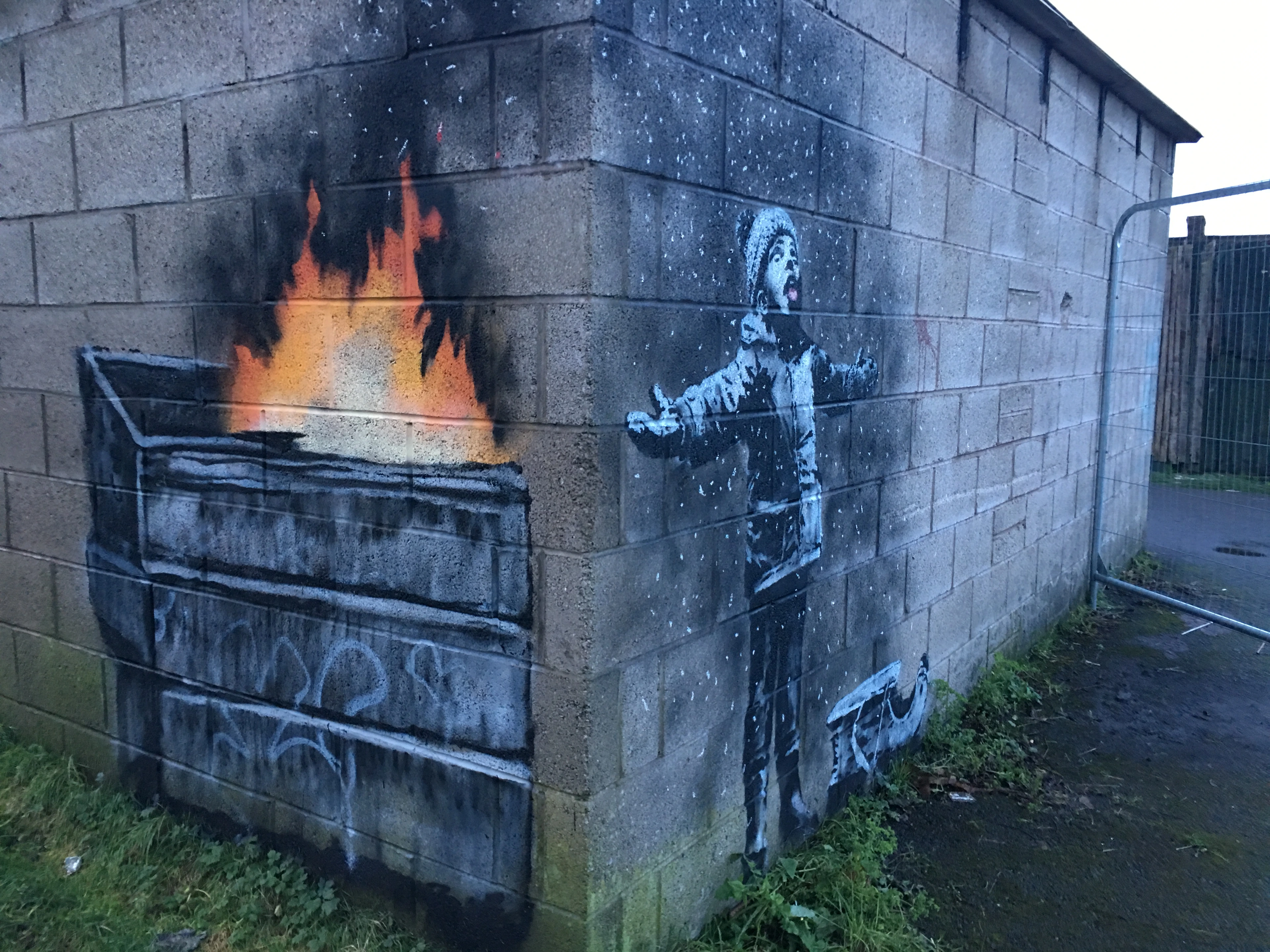 Banksy's 'Season's Greetings' mural on a concrete wall, showing a child with outstretched arms catching ash from a burning dumpster, mistaking it for snow.
