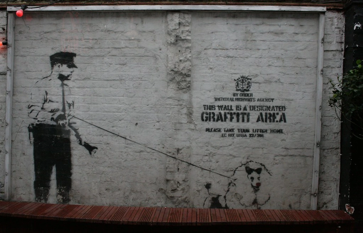 Banksy's 'Designated Graffiti Area' artwork featuring a police officer walking a poodle with a red nose, painted on a white brick wall with official-looking text.