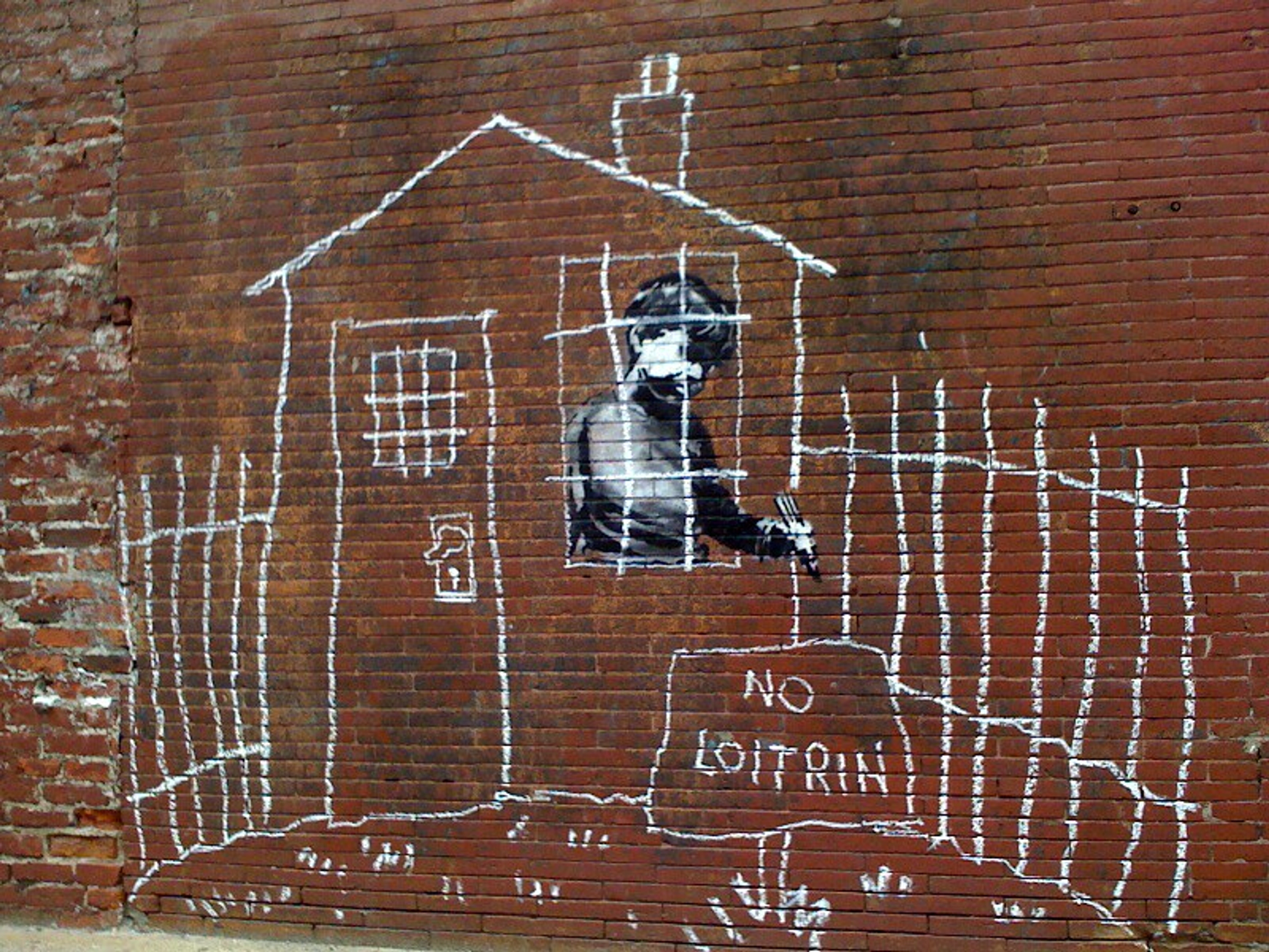 Banksy's 'NO LOITRIN' street art on a brick wall in Boston, depicting a child drawing a house with prison bars and a fence, with the text 'NO LOITRIN' below.