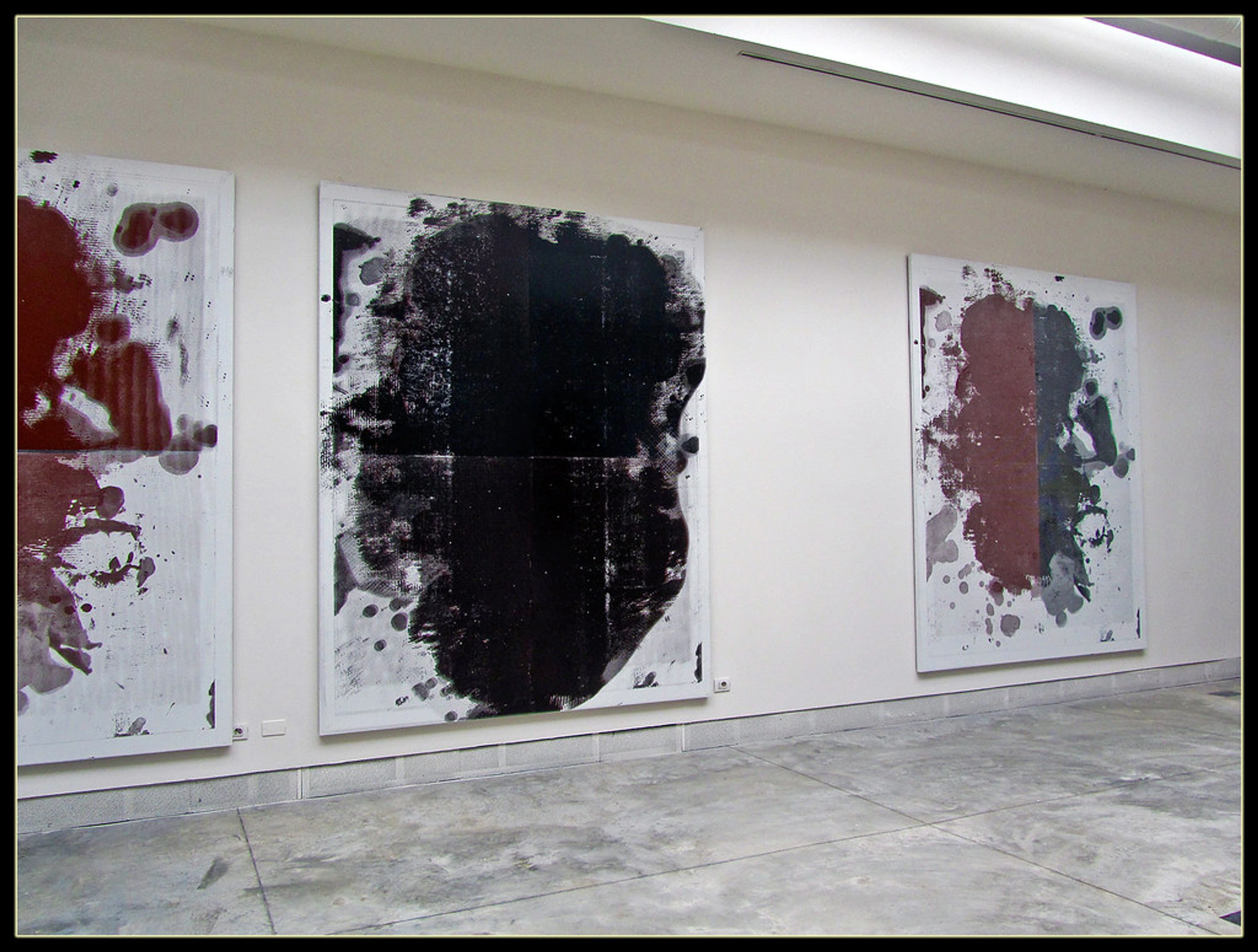 Three large abstract paintings by Christopher Wool, featuring black, dark red, and grey paint on white canvases, displayed in a modern art gallery.