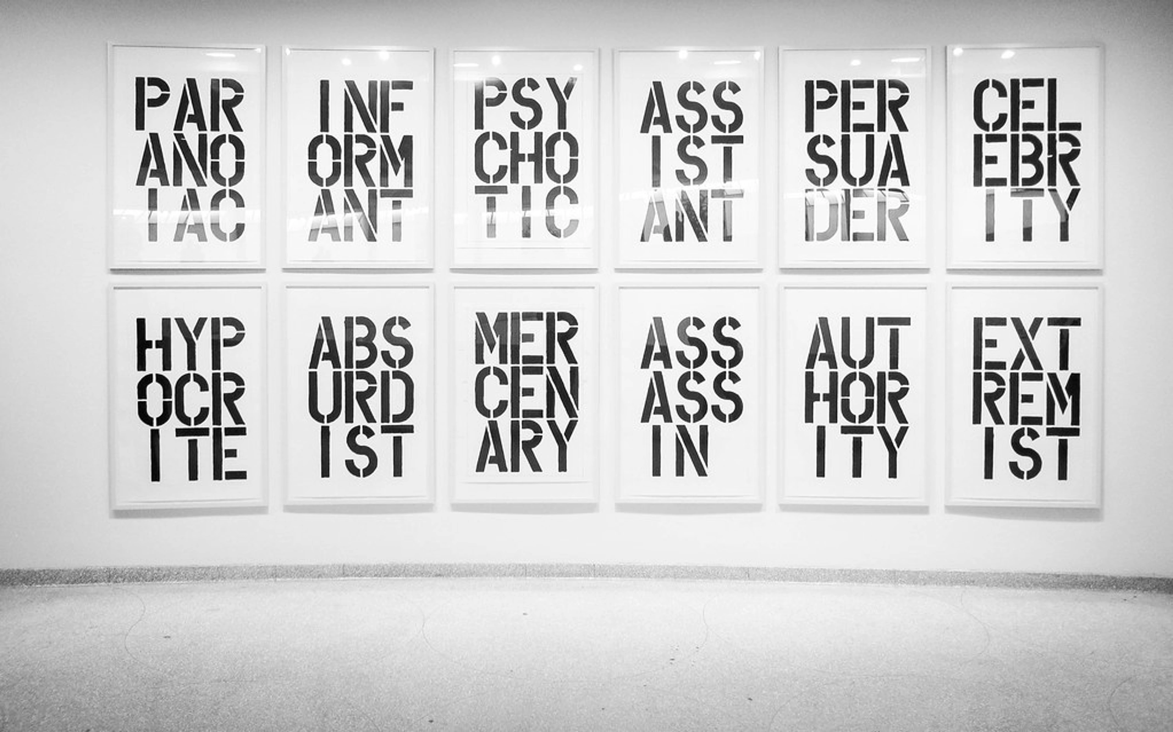 Twelve framed black and white stencil word art pieces by Christopher Wool from his 'Black Book Drawings' series, including words like 'PARANOIAC', 'INFORMANT', 'PSYCHOTIC', and 'ASSASSIN', displayed on a white wall.