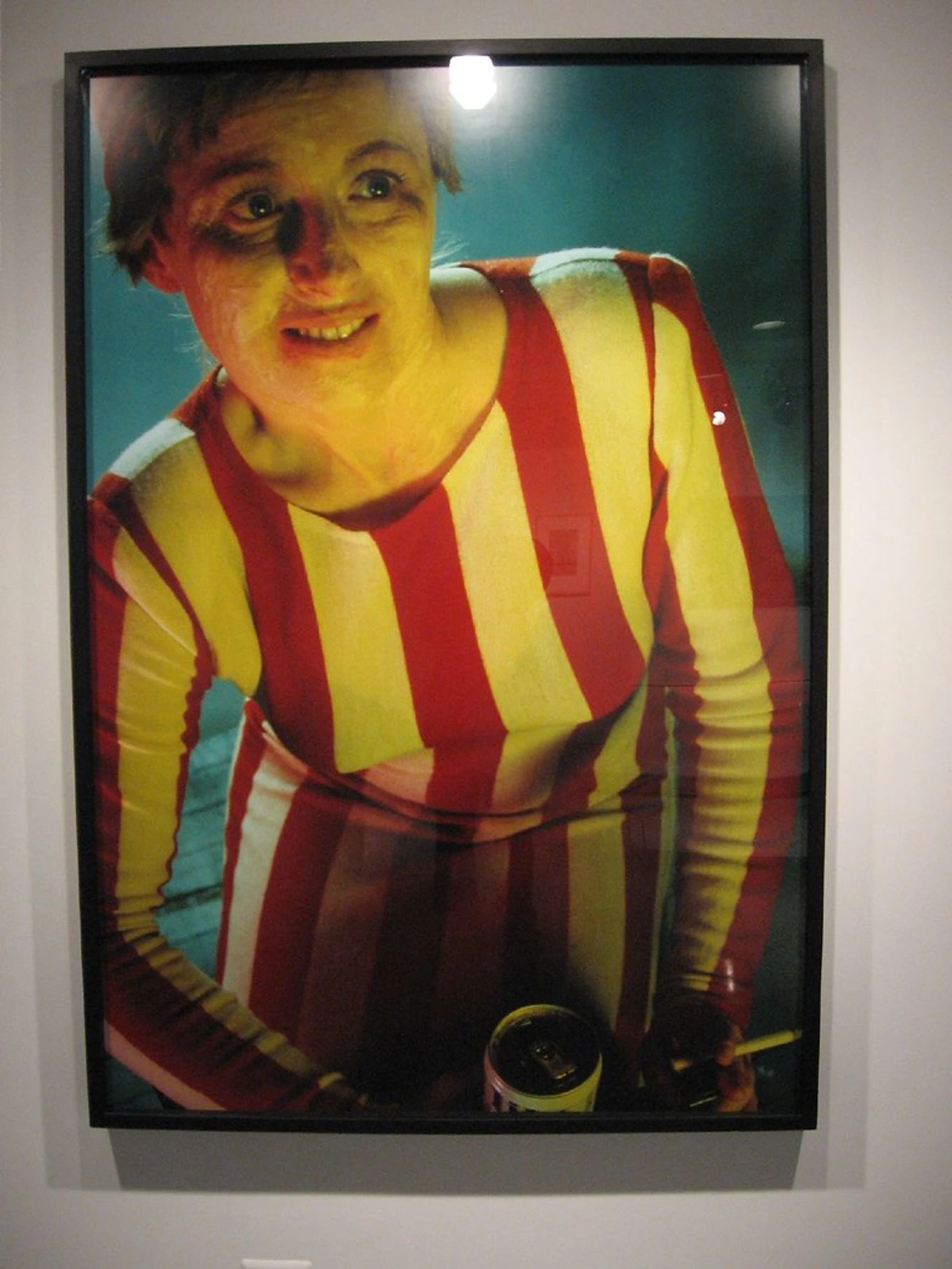 Framed color photograph by Cindy Sherman, Untitled #132 (1984), depicting a woman in a red and yellow striped outfit with a pale, smiling face. A soda can and cigarette are visible in the foreground.