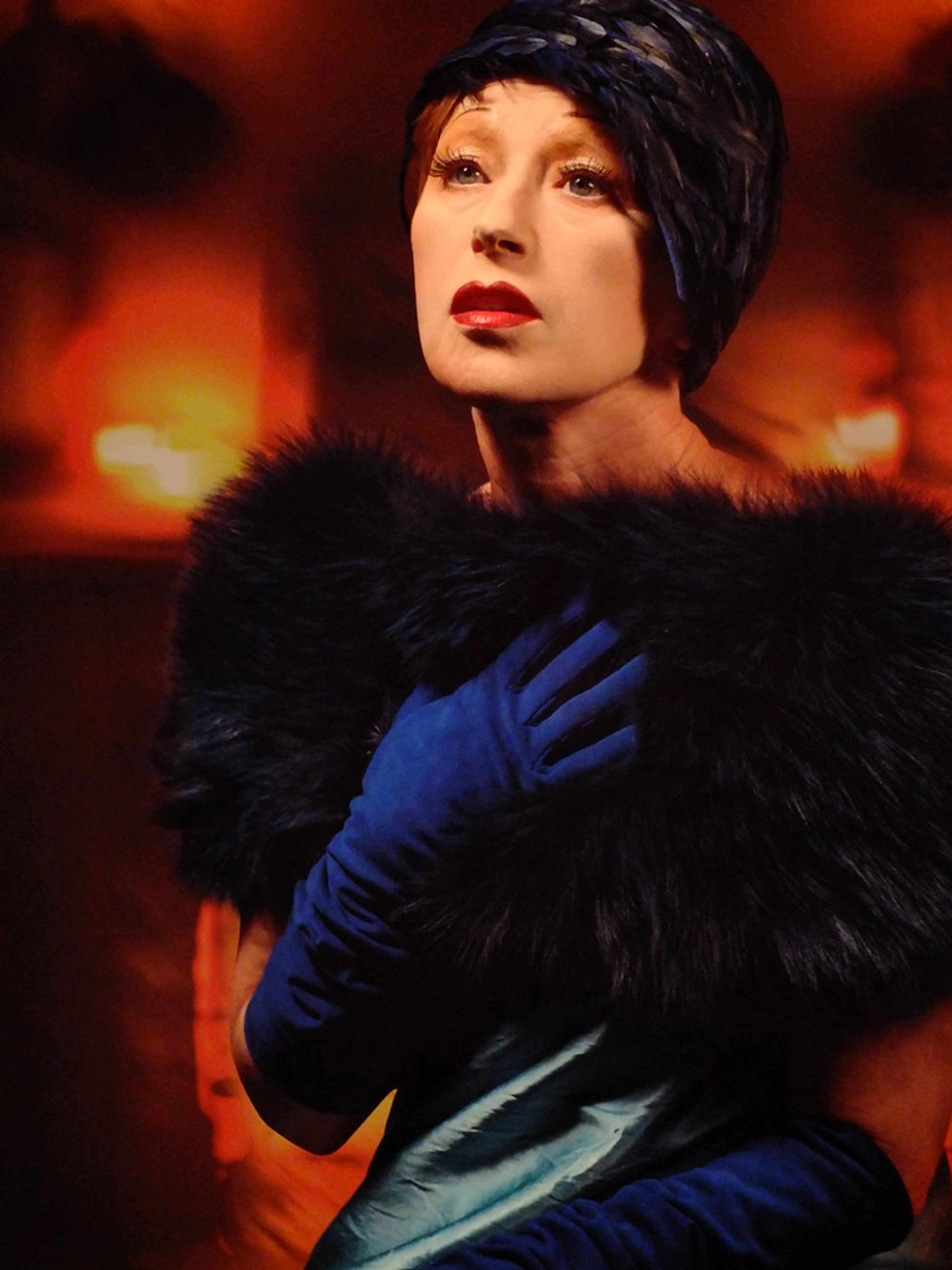 Cindy Sherman's 'Untitled #574 (2016)' self-portrait. The artist wears a blue feathered hat, red lipstick, dark fur stole, and blue gloves, looking up against a warm, blurry background.
