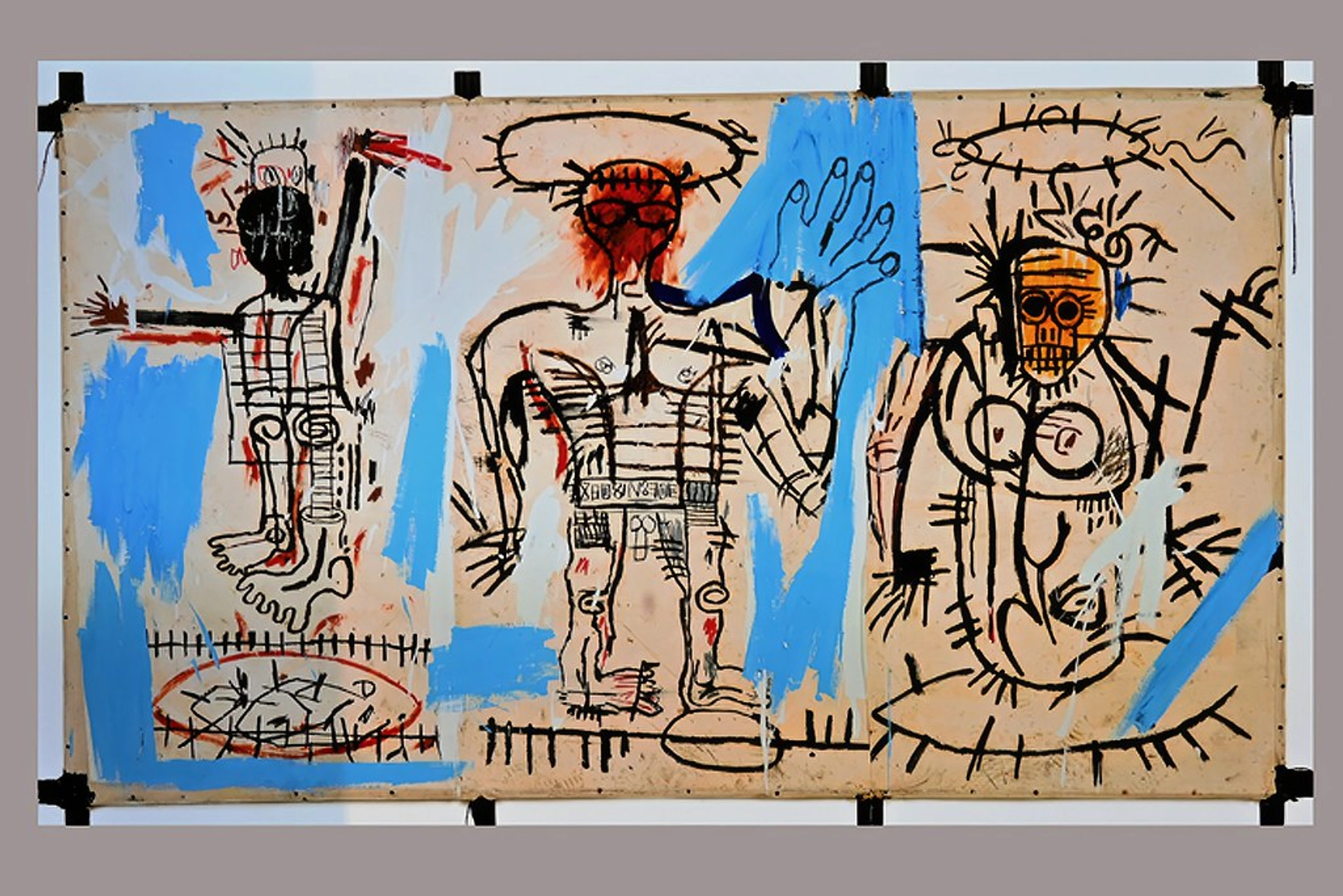 A large, multi-panel neo-expressionist painting by Jean-Michel Basquiat featuring three stylized, skeletal figures with black outlines, blue paint splashes, and red/orange accents on a beige background. The figures have distinct heads, one black, one red, and one orange skull-like.