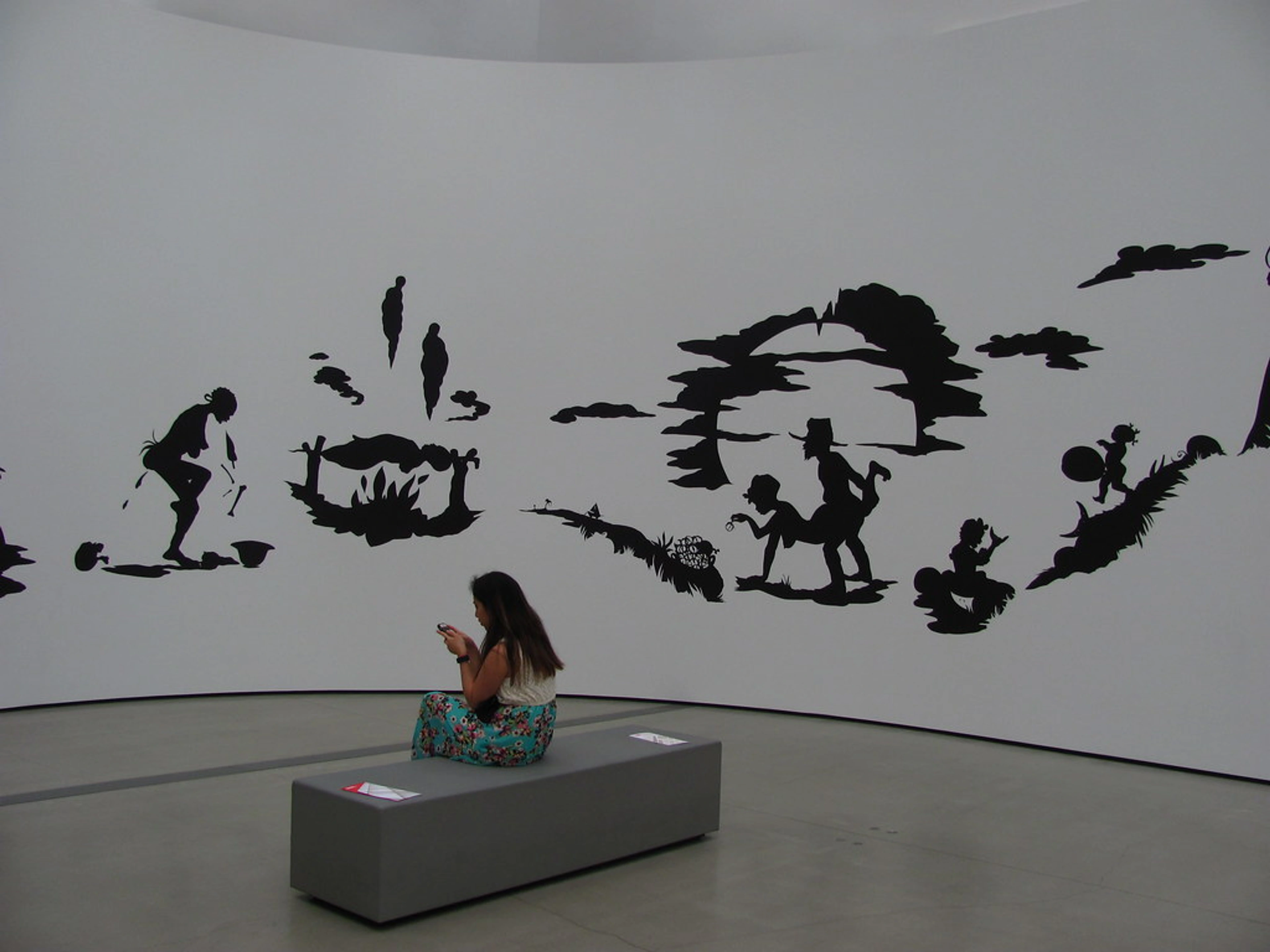 A visitor views Kara Walker's 'African't' silhouette art installation on a large curved wall at The Broad Museum. The black cutouts depict various scenes, with a woman sitting on a bench in the foreground.